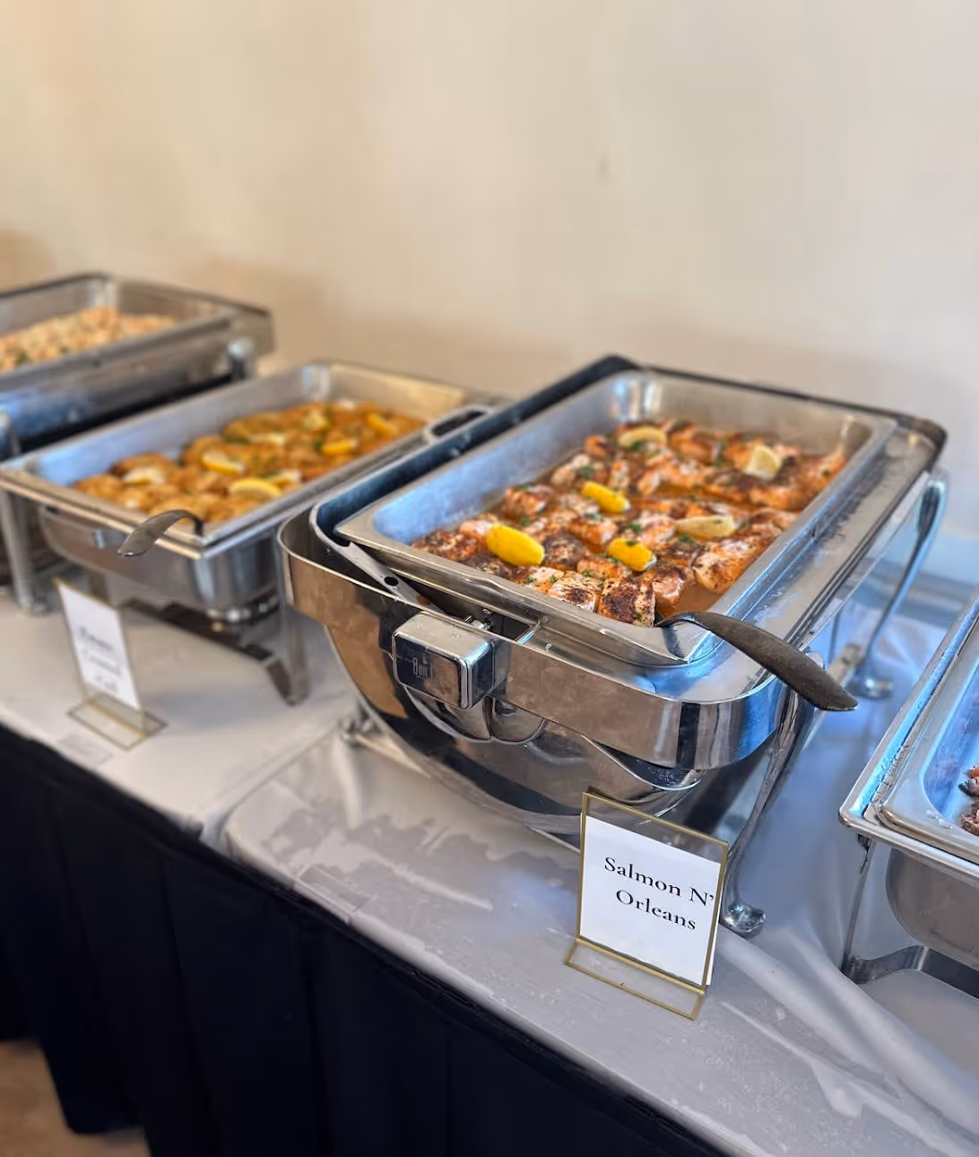 Buffet setup with trays of cooked food including a tray of salmon garnished with lemon wedges labeled 'Salmon N' Orleans', a Tangier Catering favorite.