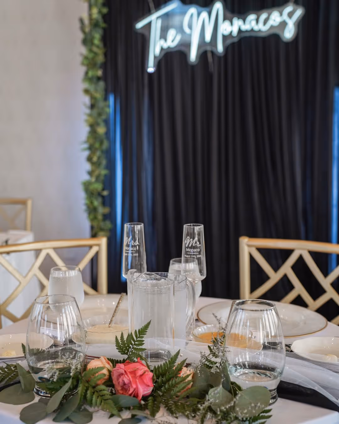 Wedding reception table set with floral centerpiece, glassware including 'Mr.' and 'Mrs.' champagne flutes, and elegant plates against a backdrop.