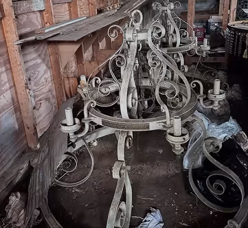 Rustic, vintage wrought iron chandelier with ornate scrollwork resting on a wooden floor inside a weathered barn. These are the old Bank chandeliers that have been repurposed.