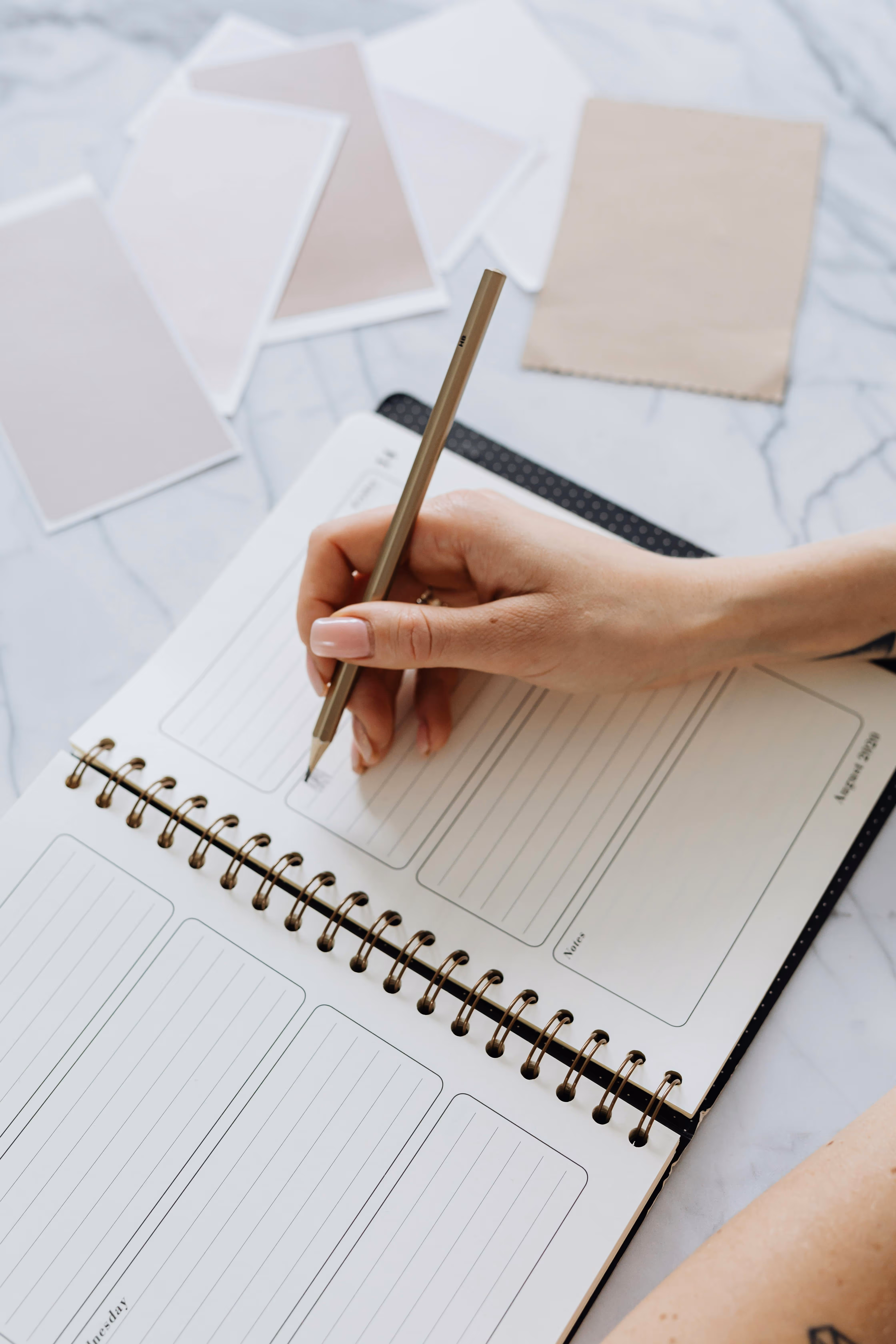 Person writing in a spiral-bound planner notebook with color swatches on a marble surface.