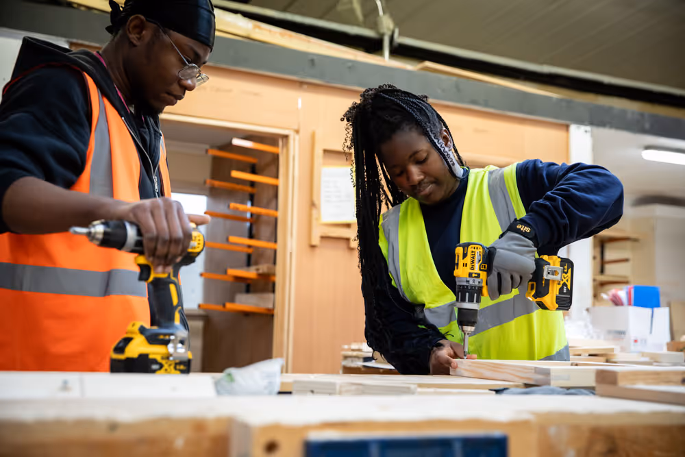 Two construction workers wearing safety vests using drills on wooden planks indoors.