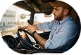 Man with beard wearing a hat and blue shirt driving a car, viewed from the passenger side.
