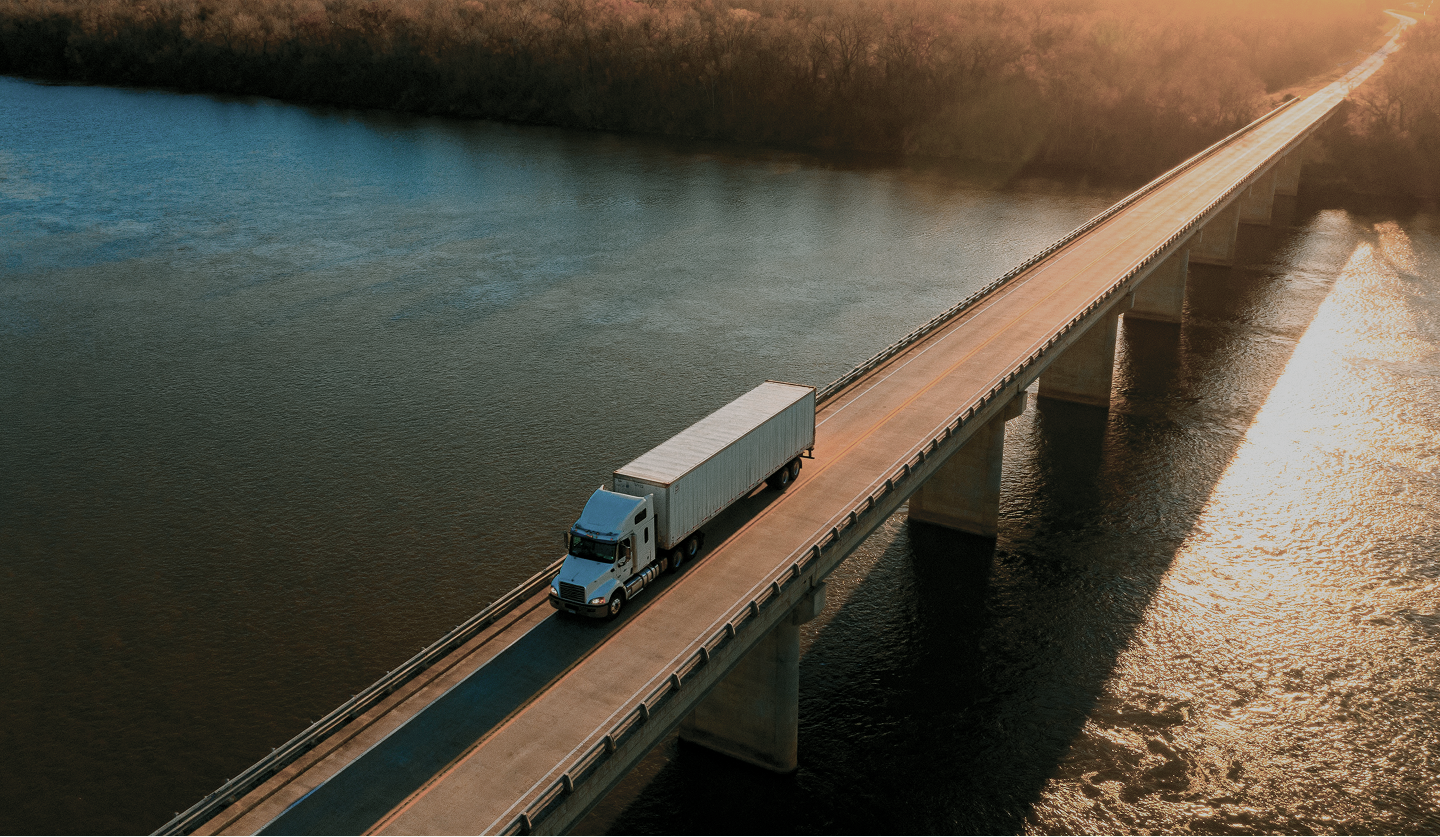 White semi-truck driving on a long bridge over a calm river during sunset.