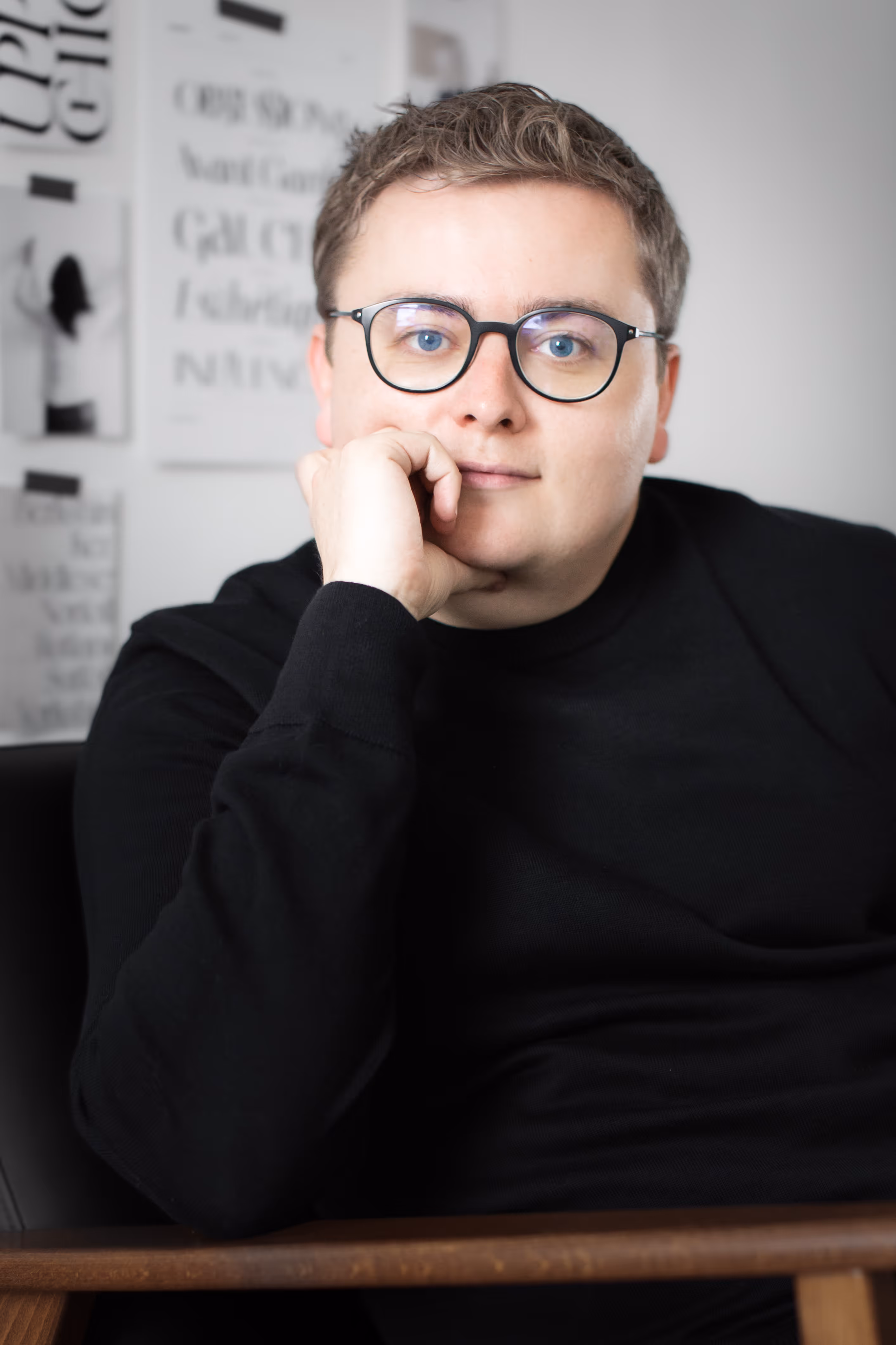 Person with short blonde hair and glasses wearing a black sweater, resting their chin on their hand and looking at the camera. Portrait of Jack Watkins.