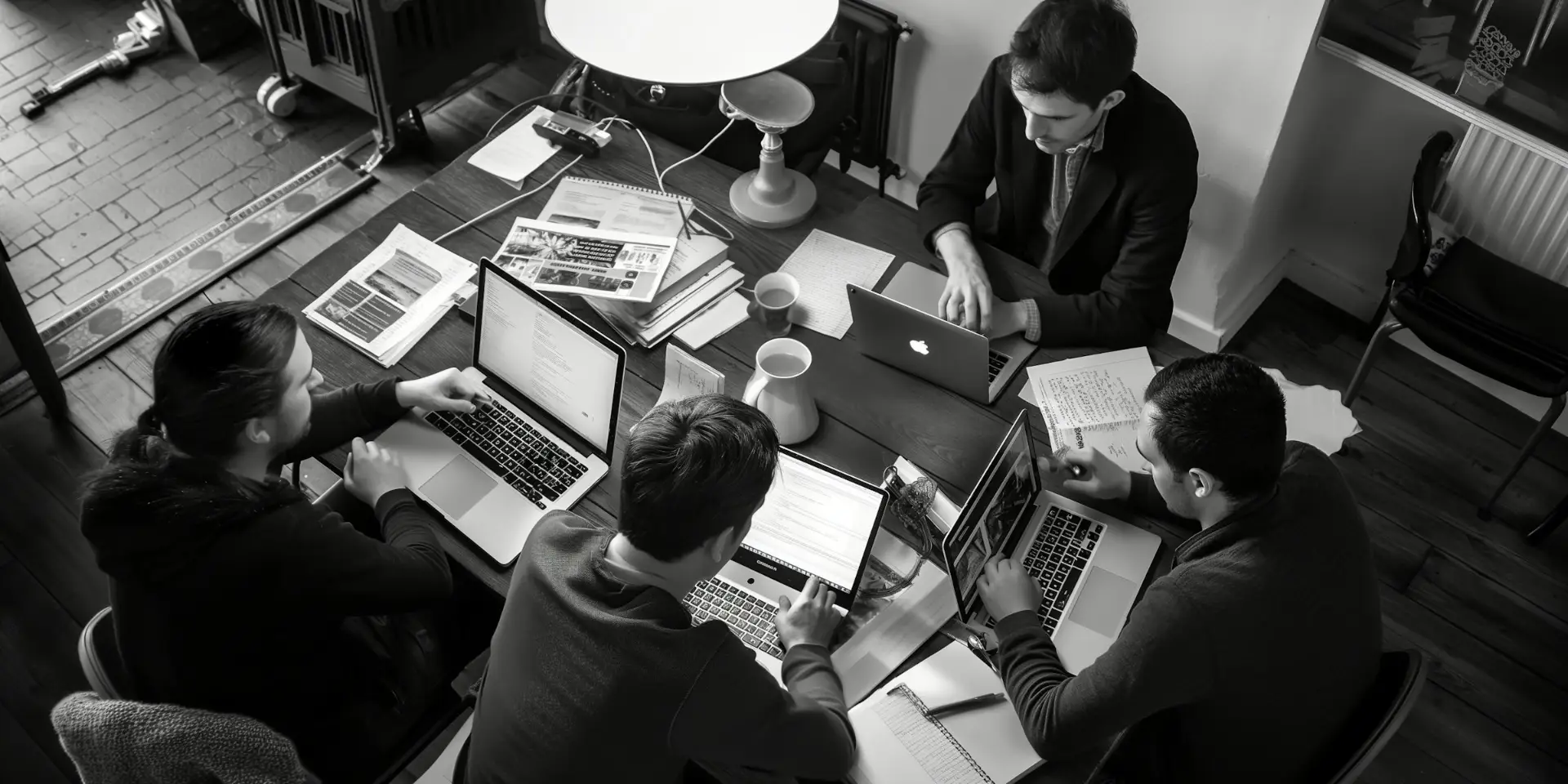 Four people working collaboratively on laptops around a wooden table with papers and cups.