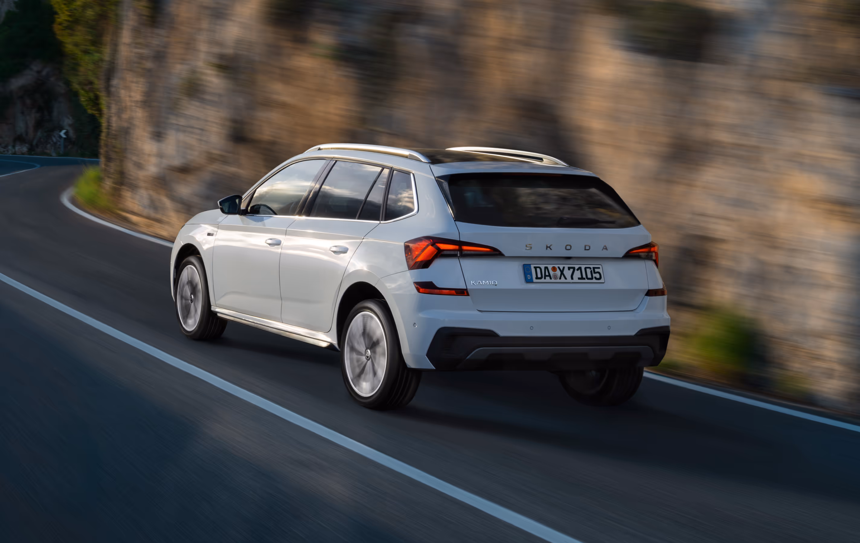 Dynamische Heck-Schrägansicht eines Škoda Kamiq Balance in der Lackierung Moon-Weiß Perleffekt auf einer Landstraße. Das Bild zeigt das SUV in Fahrt vor einer Felswand und hebt die Dachreling hervor, in der sich die Sonne spiegelt. Zu sehen sind zudem die markanten LED-Heckleuchten sowie der Škoda Schriftzug auf der Heckklappe.