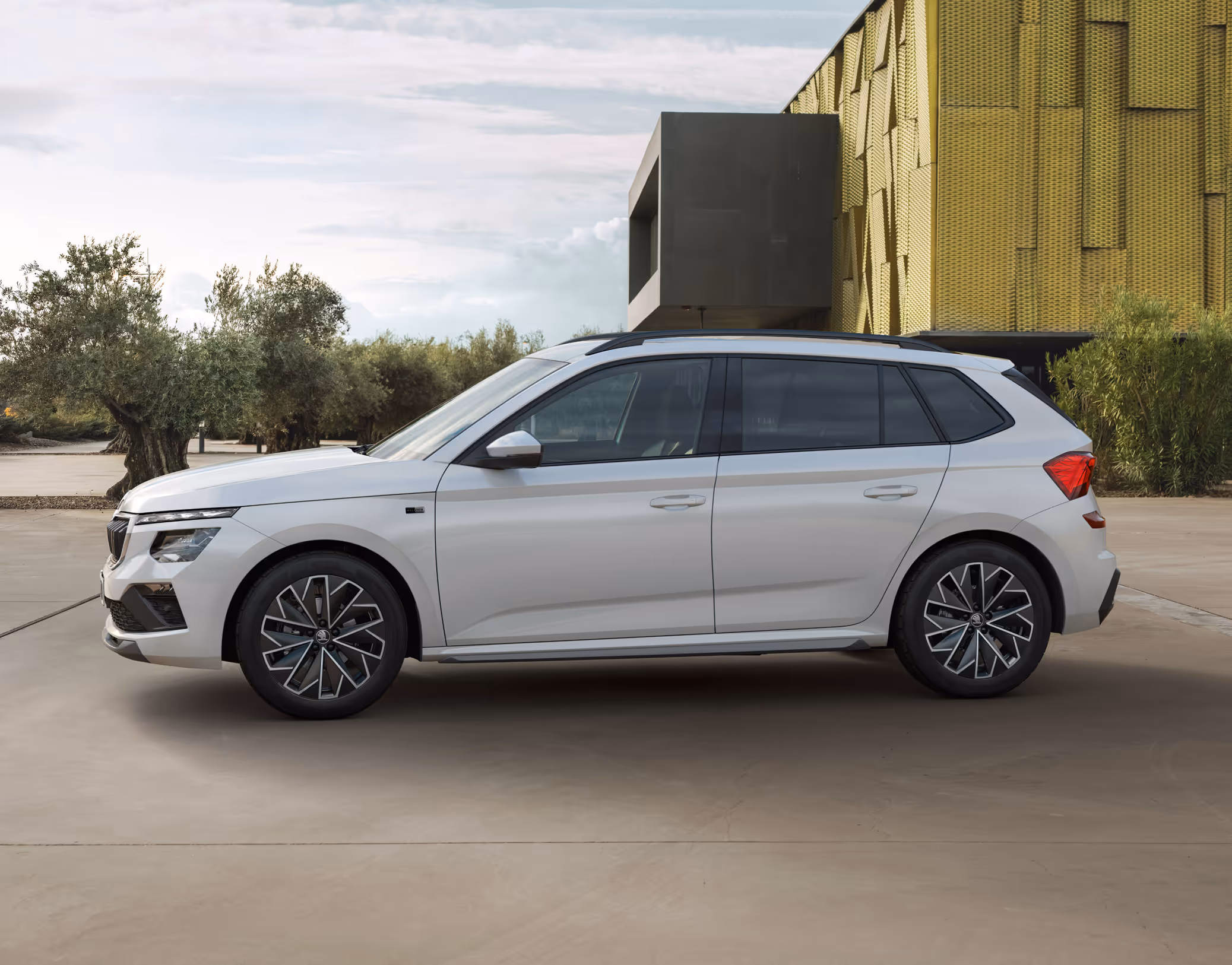 Ein Škoda Kamiq Balance in der Lackierung Moon-Weiß Perleffekt ist in der Seitenansicht vor einer modernen Architektur geparkt. Das Exterieur-Design des Crossovers wird durch die Kombination aus urbaner und natürlicher Umgebung hervorgehoben.