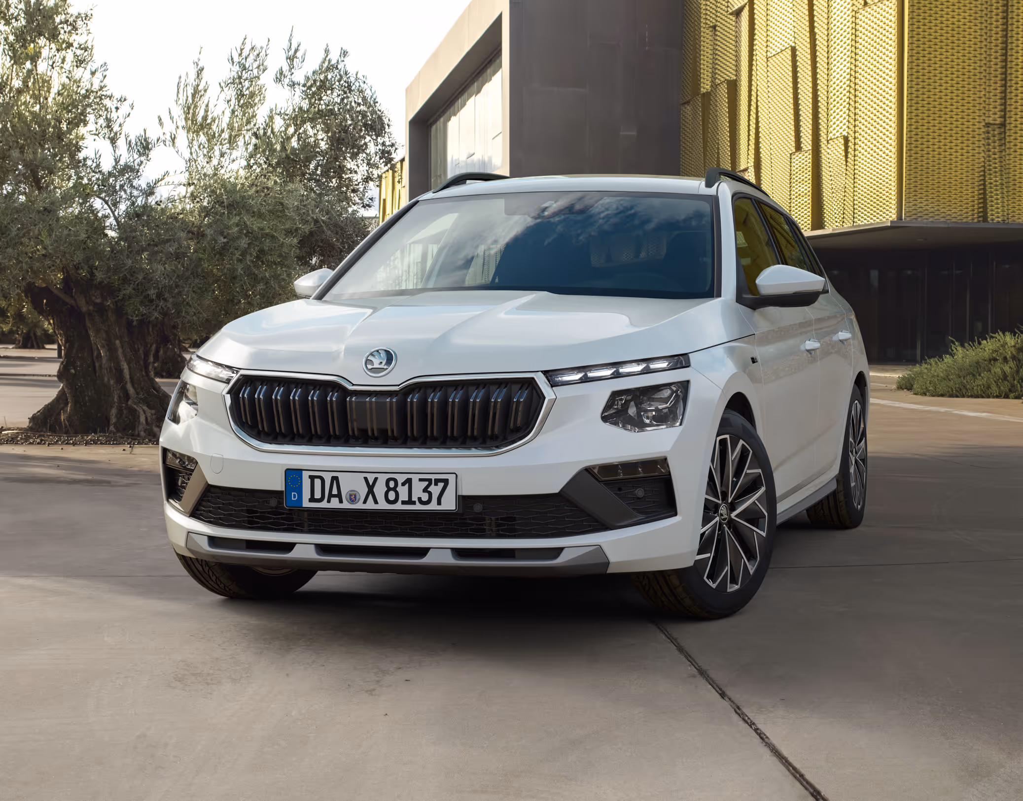 Ein Škoda Kamiq Balance in der Lackierung Moon-Weiß Perleffekt ist in einer Dreiviertelansicht von vorne auf einem Vorplatz geparkt. Das Fahrzeug steht neben einem Olivenbaum vor einem modernen Gebäude mit goldener Fassade.