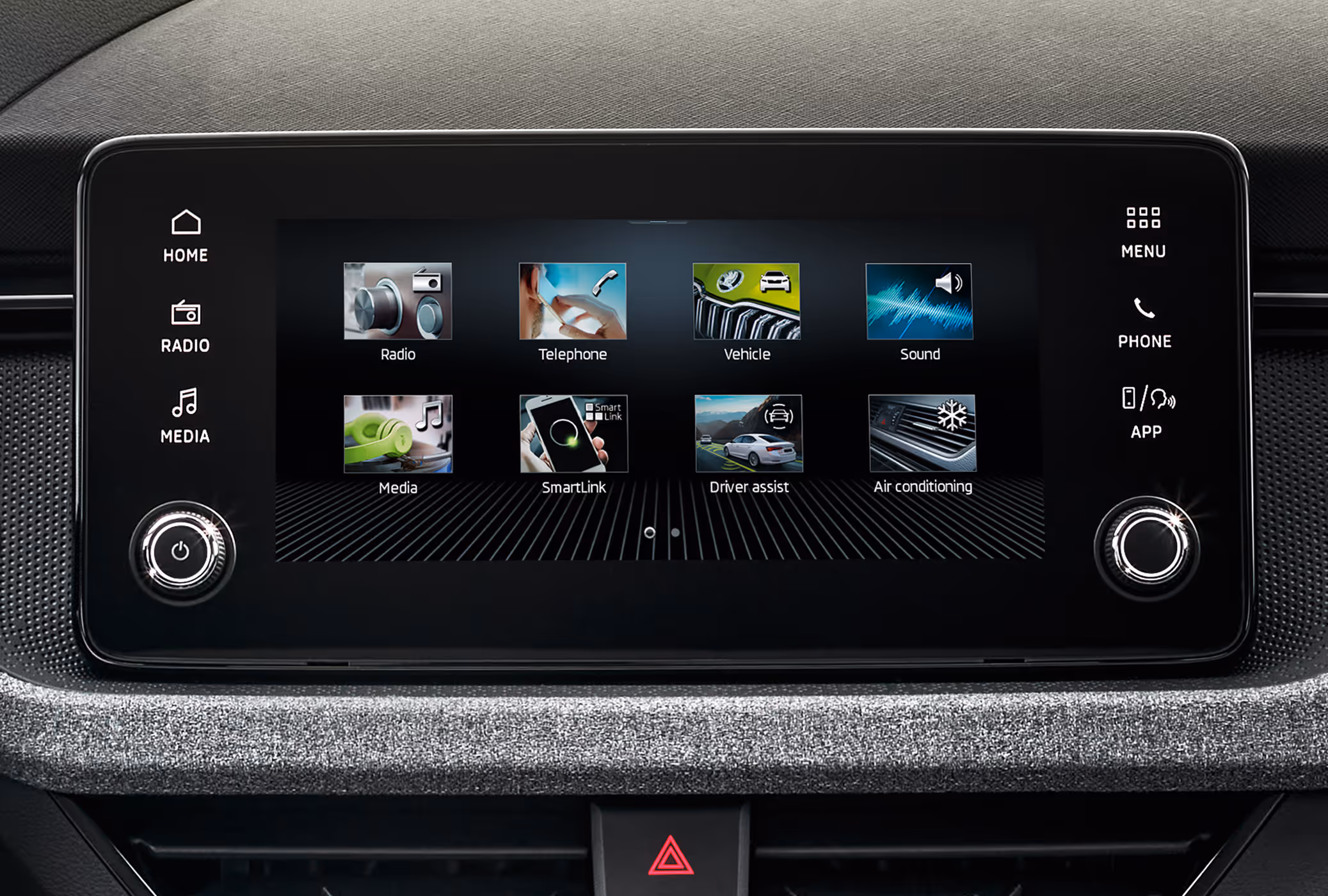 Nahaufnahme des Infotainment-Displays im Škoda Kamiq Balance, das im Hauptmenü verschiedene Funktionskacheln wie Radio, SmartLink und Klimaanlage anzeigt. Der Touchscreen wird von seitlichen Sensortasten sowie zwei physischen Drehreglern eingerahmt. Unterhalb des Bildschirms ist die für die Ausstattungslinie typische Dekorleiste mit grauem Stoffbezug zu sehen.