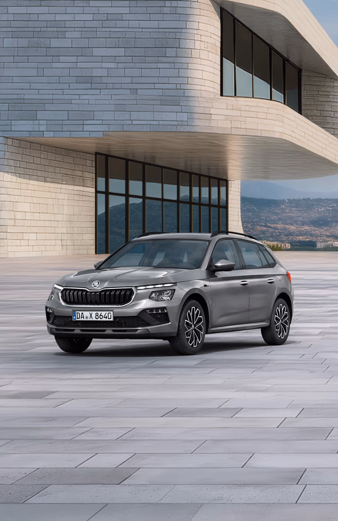 Front-Schrägansicht eines Škoda Kamiq Balance in der Lackierung Graphite-Grau Metallic vor einer modernen Gebäudekulisse. Das SUV ist mit einer schwarzen Dachreling und dunklen, glanzgedrehten Leichtmetallfelgen ausgestattet. Ein grüner Störer im unteren Bildbereich nennt einen Preisvorteil von 2.000 € sowie die inkludierte 5-Jahres-Garantie.