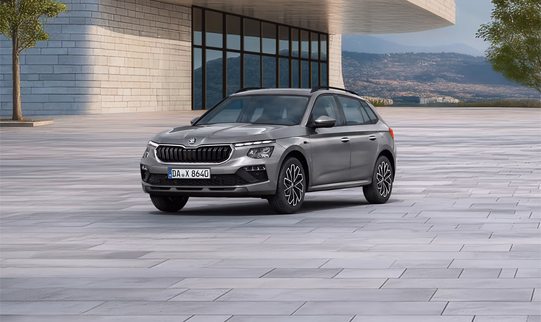 Front-Schrägansicht eines Škoda Kamiq Balance in der Lackierung Graphite-Grau Metallic vor einer modernen Gebäudekulisse. Das SUV ist mit einer schwarzen Dachreling und dunklen, glanzgedrehten Leichtmetallfelgen ausgestattet. Ein grüner Störer im unteren Bildbereich nennt einen Preisvorteil von 2.000 € sowie die inkludierte 5-Jahres-Garantie.