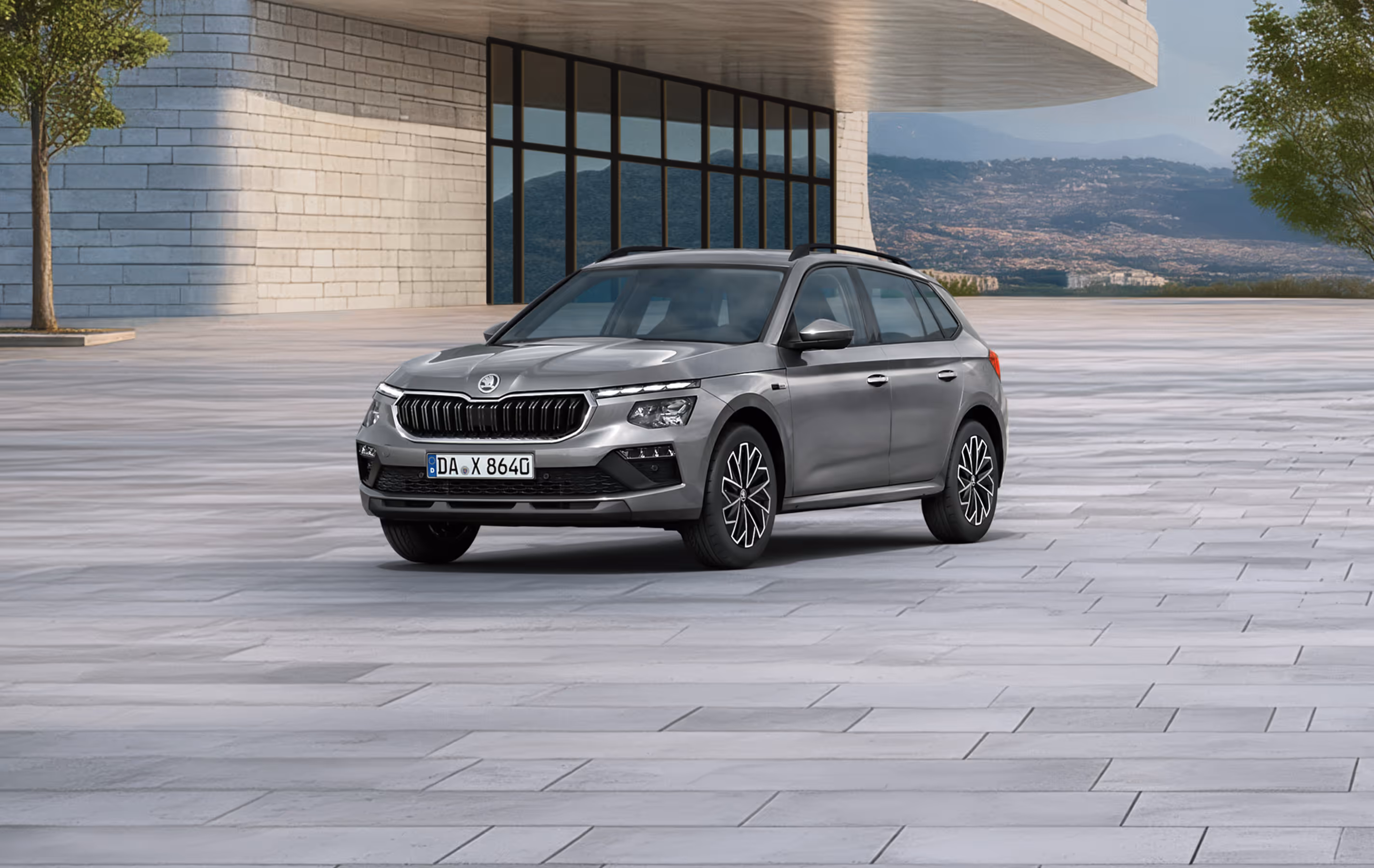 Front-Schrägansicht eines Škoda Kamiq Balance in der Lackierung Graphite-Grau Metallic vor einer modernen Gebäudekulisse. Das SUV ist mit einer schwarzen Dachreling und dunklen, glanzgedrehten Leichtmetallfelgen ausgestattet. Ein grüner Störer im unteren Bildbereich nennt einen Preisvorteil von 2.000 € sowie die inkludierte 5-Jahres-Garantie.