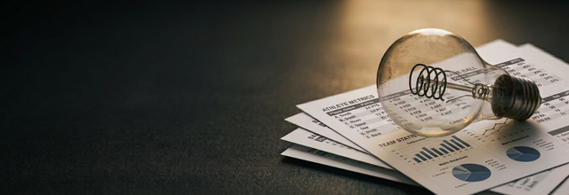 An unlit lightbulb resting on a stack of weathered stat sheets, lit by a single warm beam of light in a dark room.