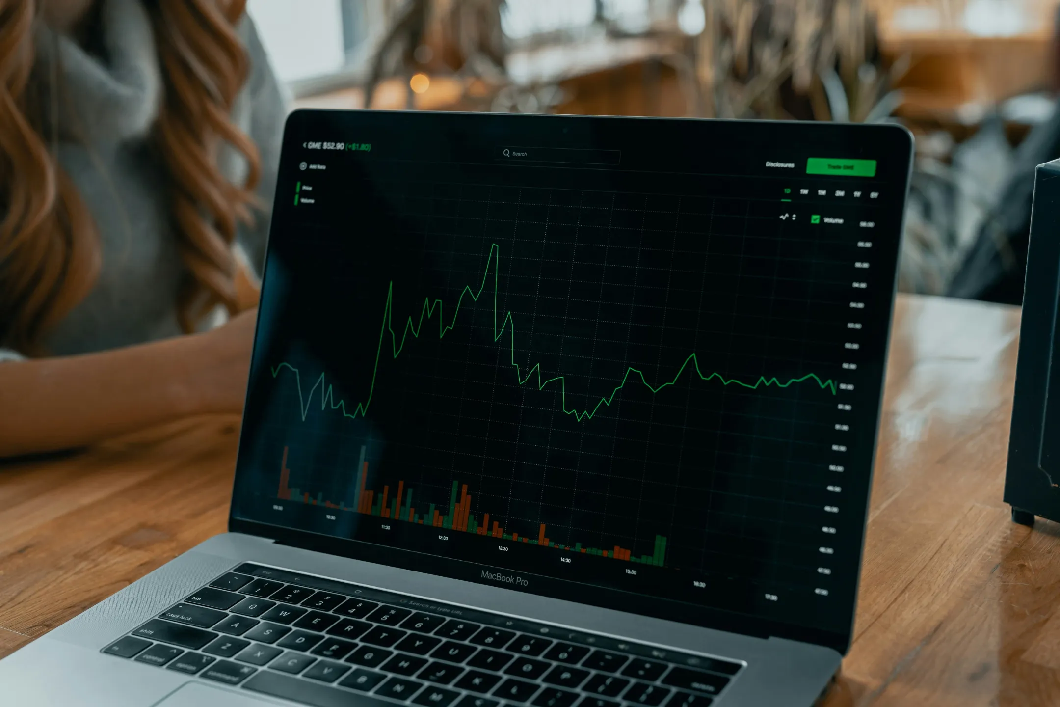 MacBook Pro laptop displaying a green line graph with stock price and volume data on screen in a wooden table setting.