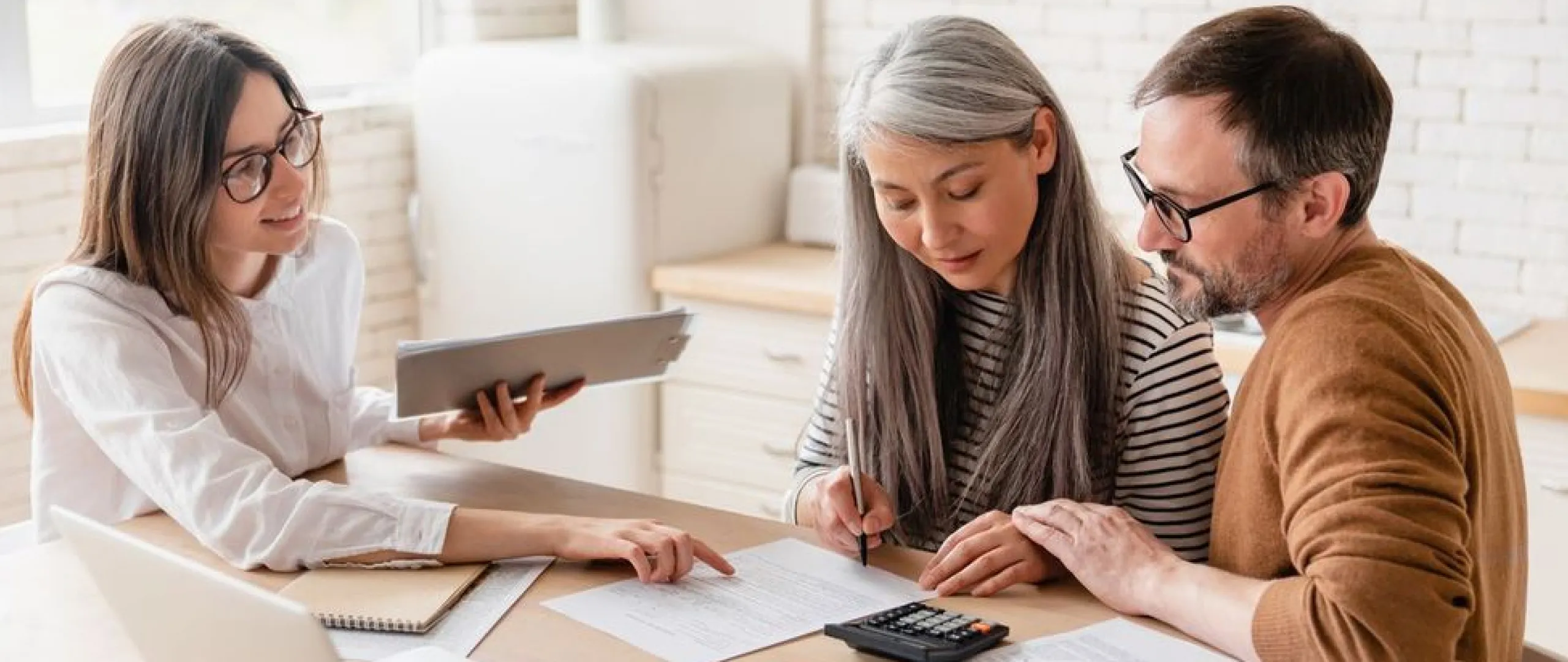 Couple reviewing documents and signing papers at a desk while a woman with glasses assists them.