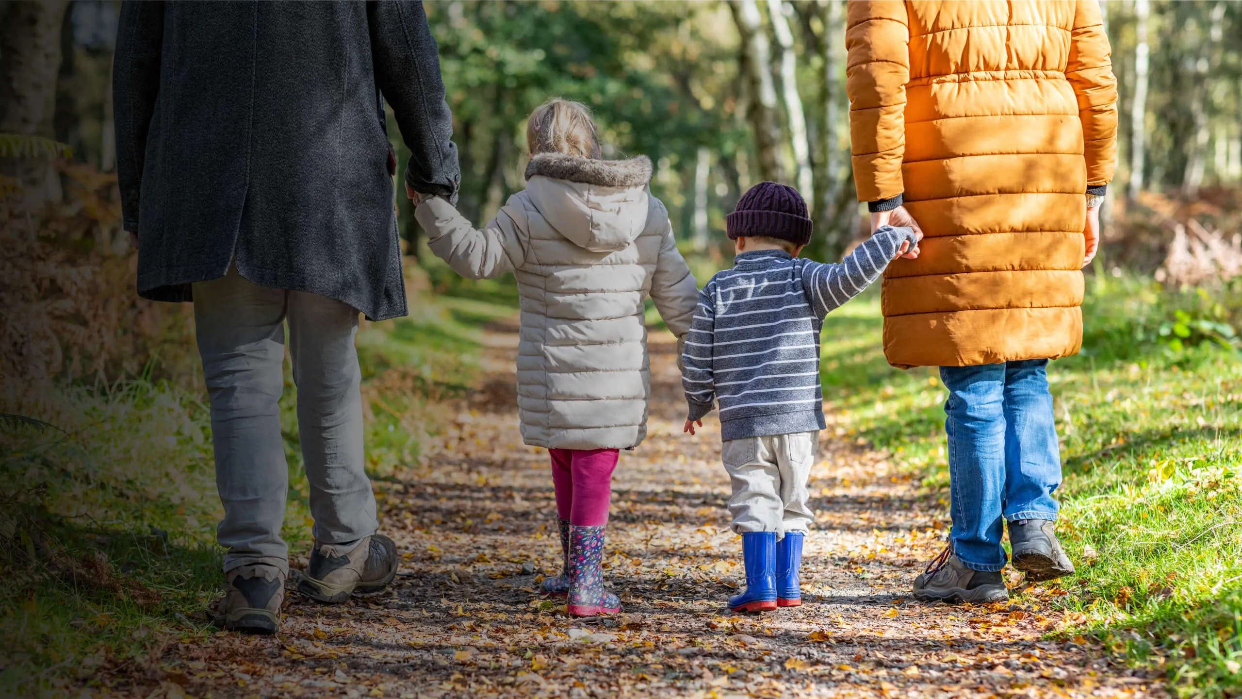 Two adults and two children walking on a leaf-covered forest path in autumn, holding hands.