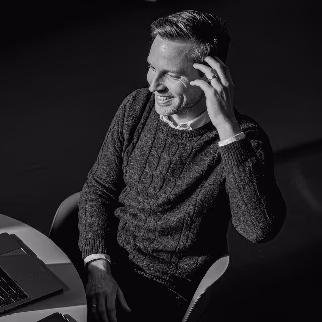 Man in a cable-knit sweater sitting at a round table with a laptop, smiling and touching his hair.
