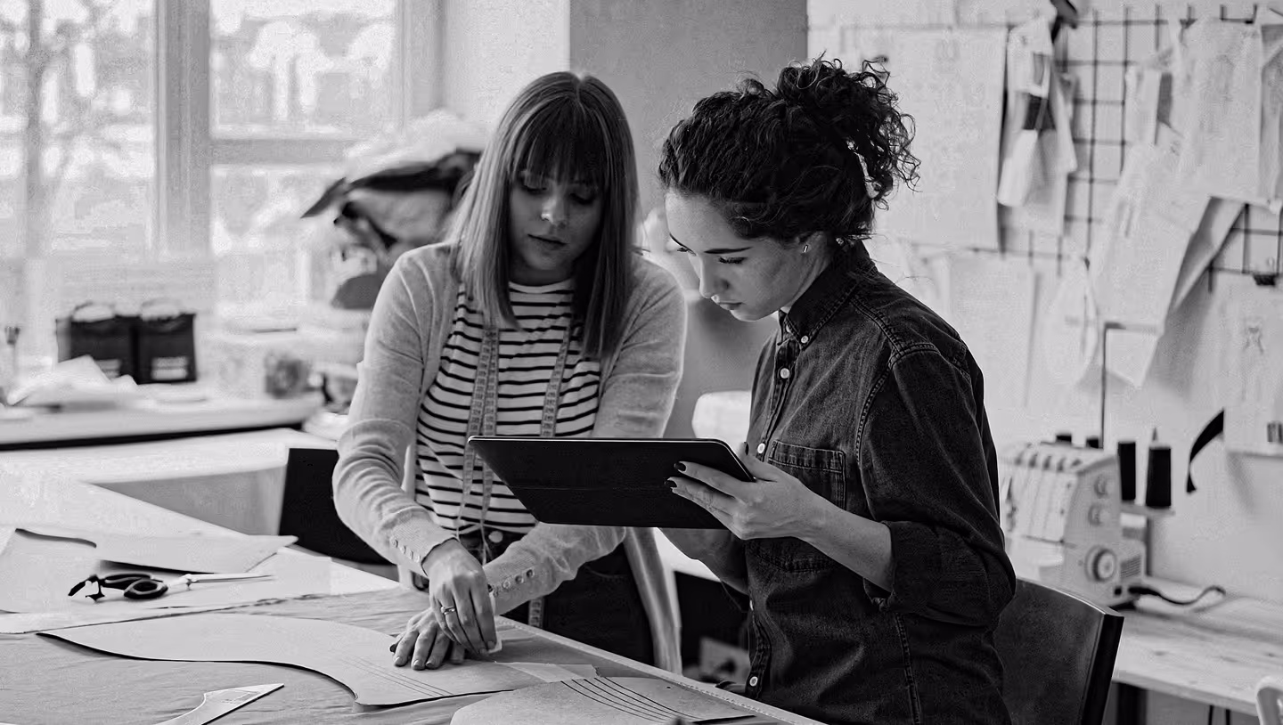 Two women working together on sewing patterns, one pointing at the fabric and the other holding a tablet in a well-lit workspace.