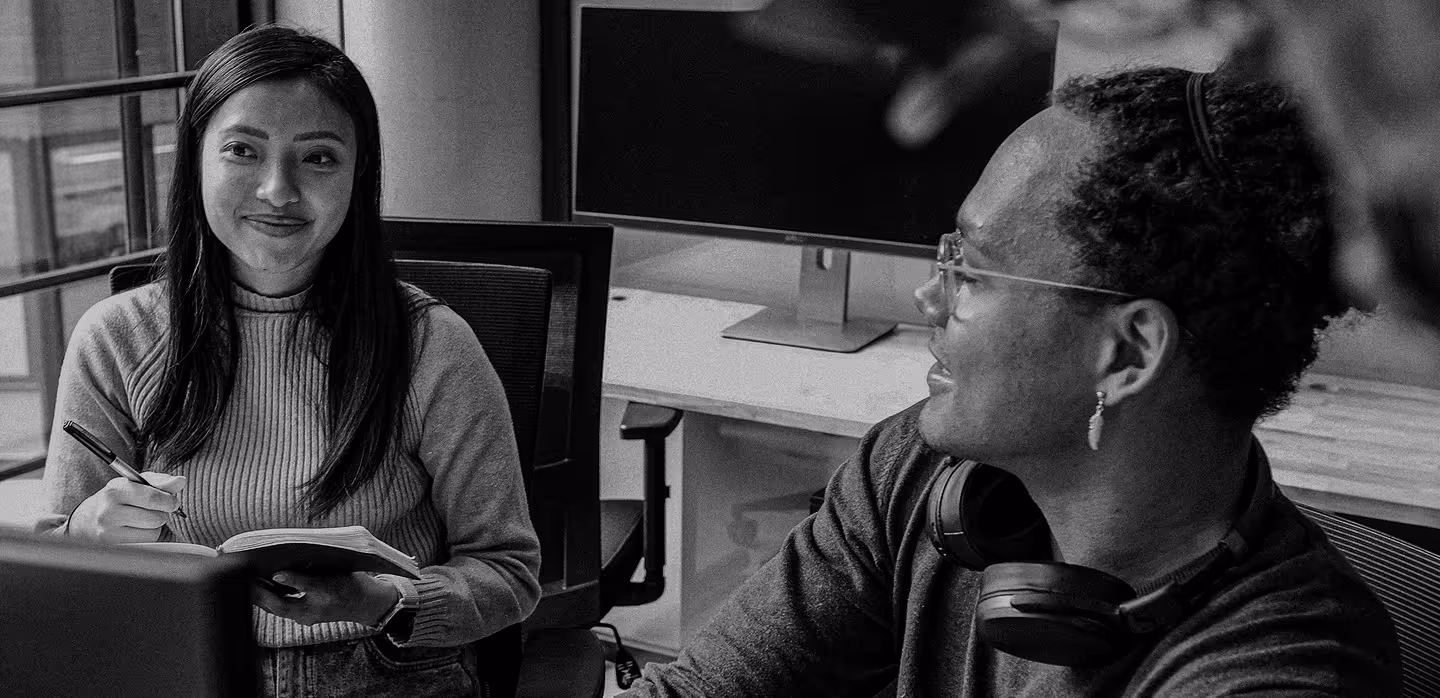 Two colleagues smiling and communicating in a modern office with a desktop computer in the background.