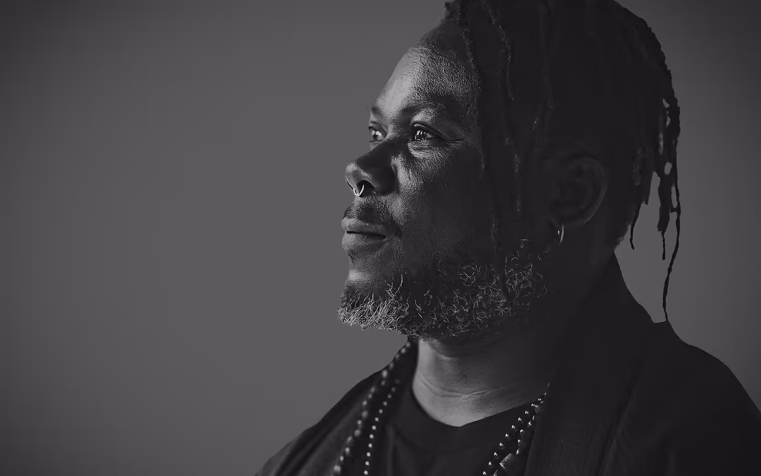 Black and white portrait of a thoughtful man with dreadlocks, a beard, and nose ring looking to the left.
