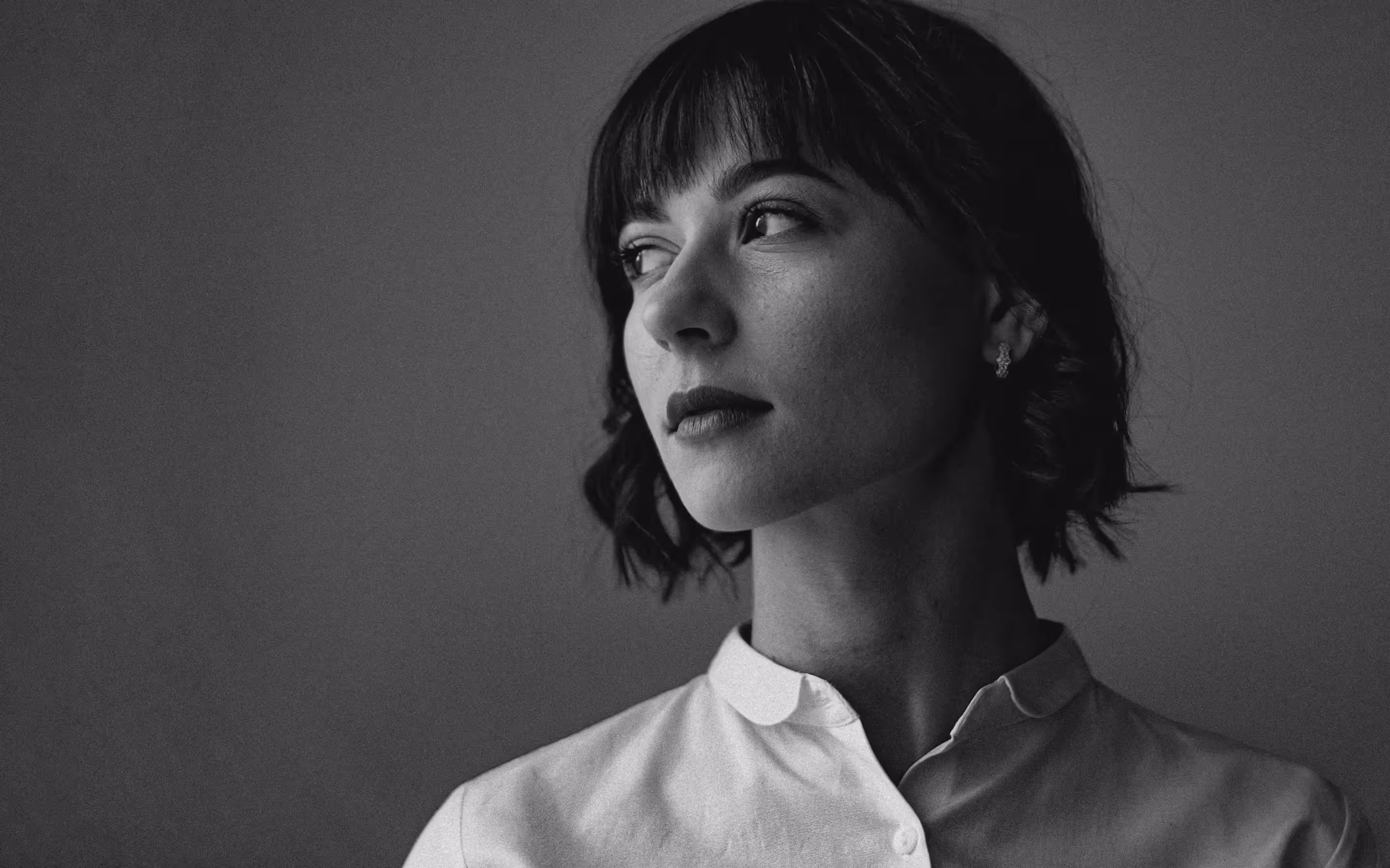 Black and white portrait of a woman with short dark hair looking away from the camera, wearing a white button-up shirt and stud earrings.