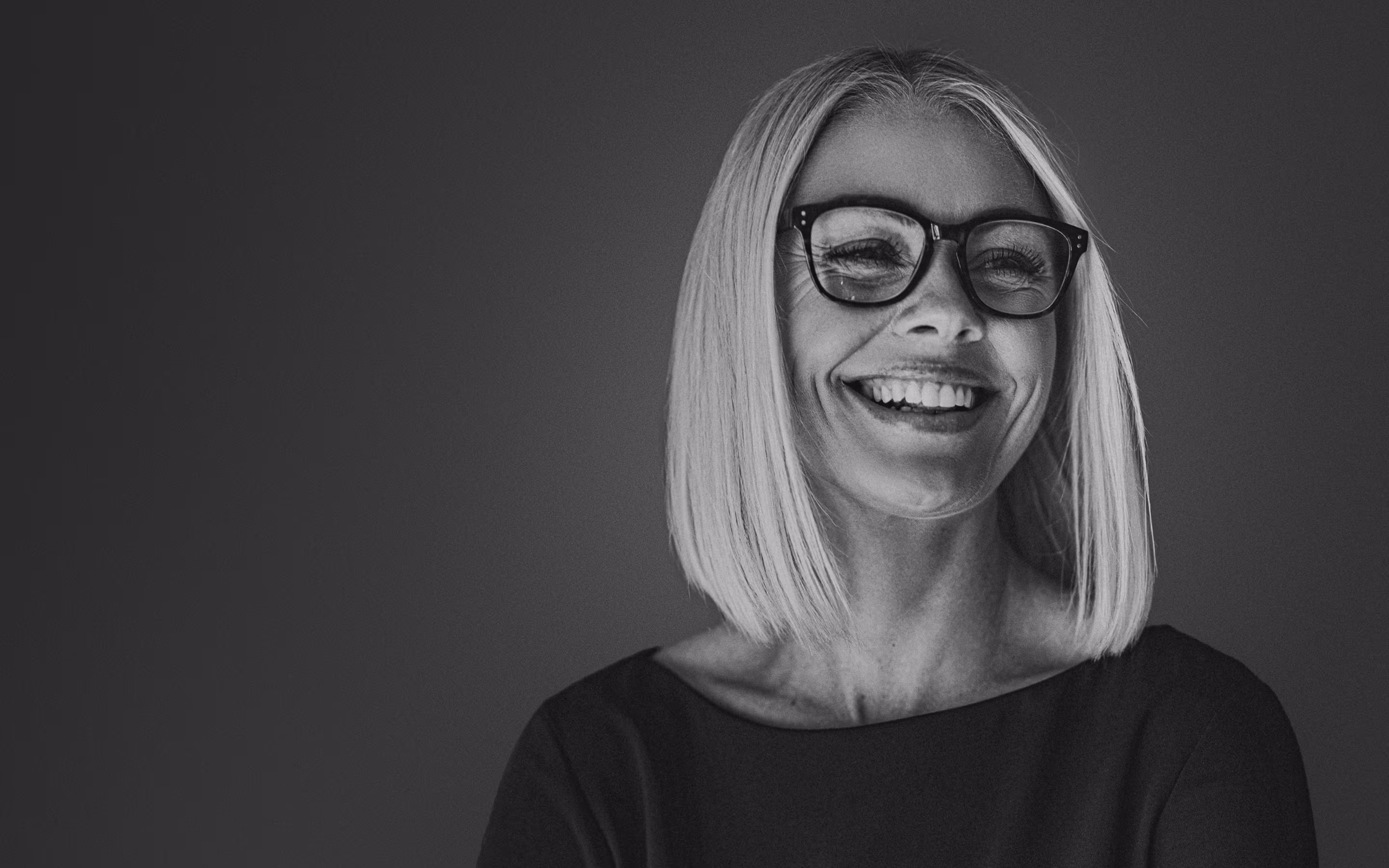Smiling middle-aged woman with straight shoulder-length blonde hair and black-rimmed glasses wearing a dark top.
