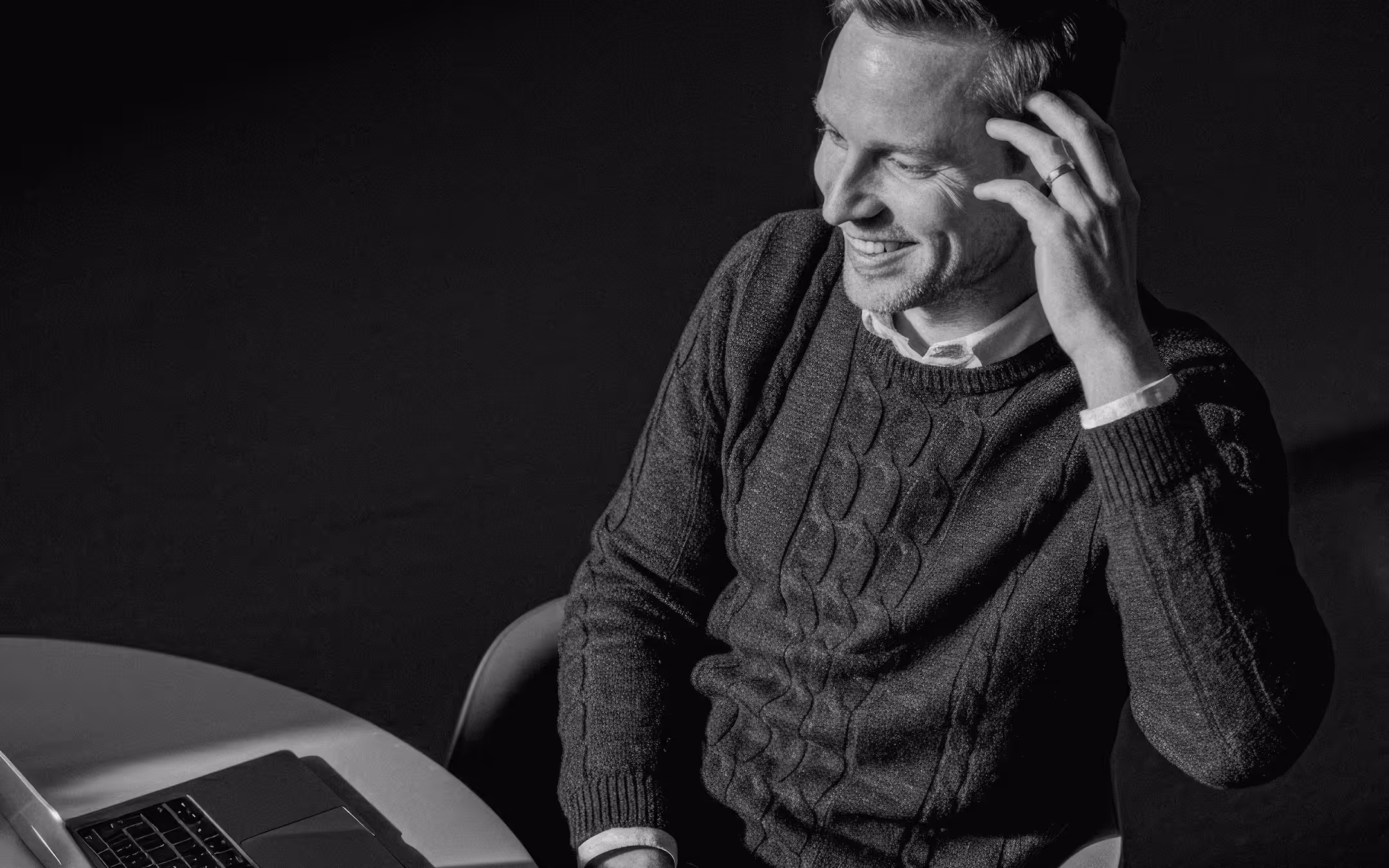 Smiling man in a cable-knit sweater sitting at a table with a laptop, touching his hair.