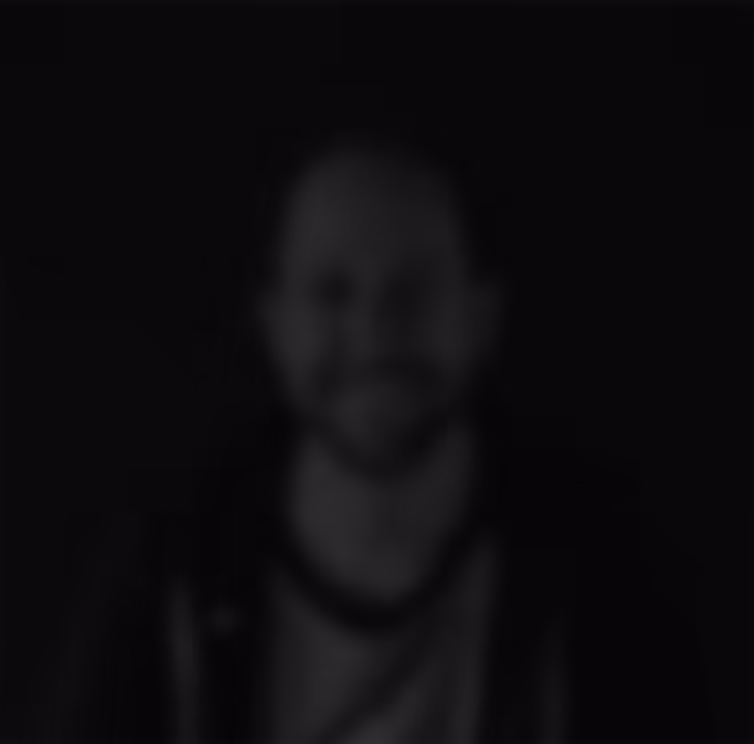 Blurry portrait of a person with short hair and a beard wearing a jacket over a t-shirt against a dark background.