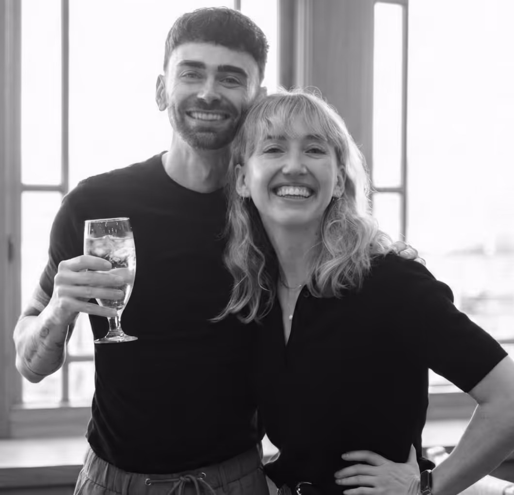 Smiling man holding a glass of iced drink and a woman with shoulder-length hair standing close together indoors.