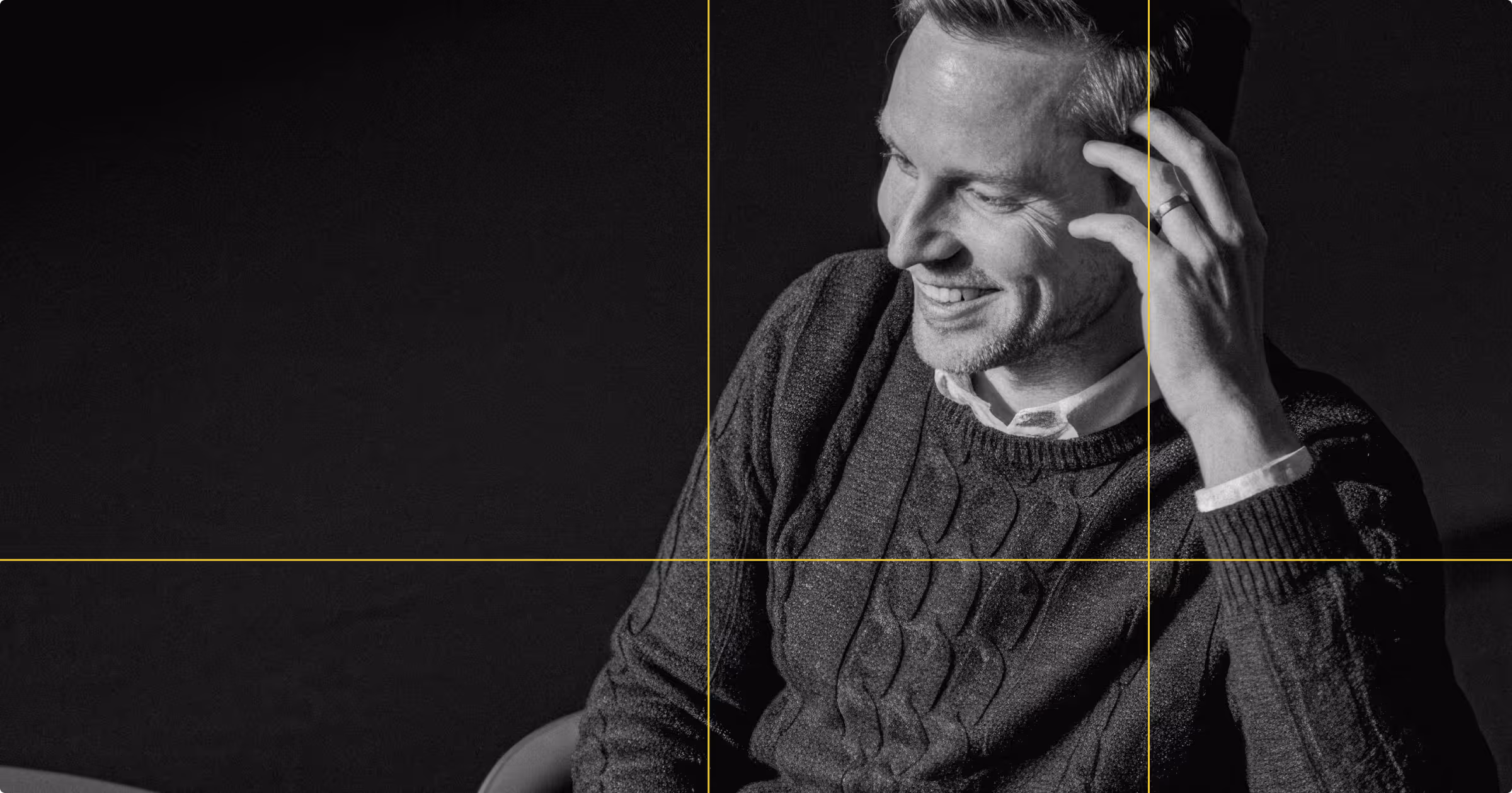 Smiling man in a cable-knit sweater touching his head against a dark background.