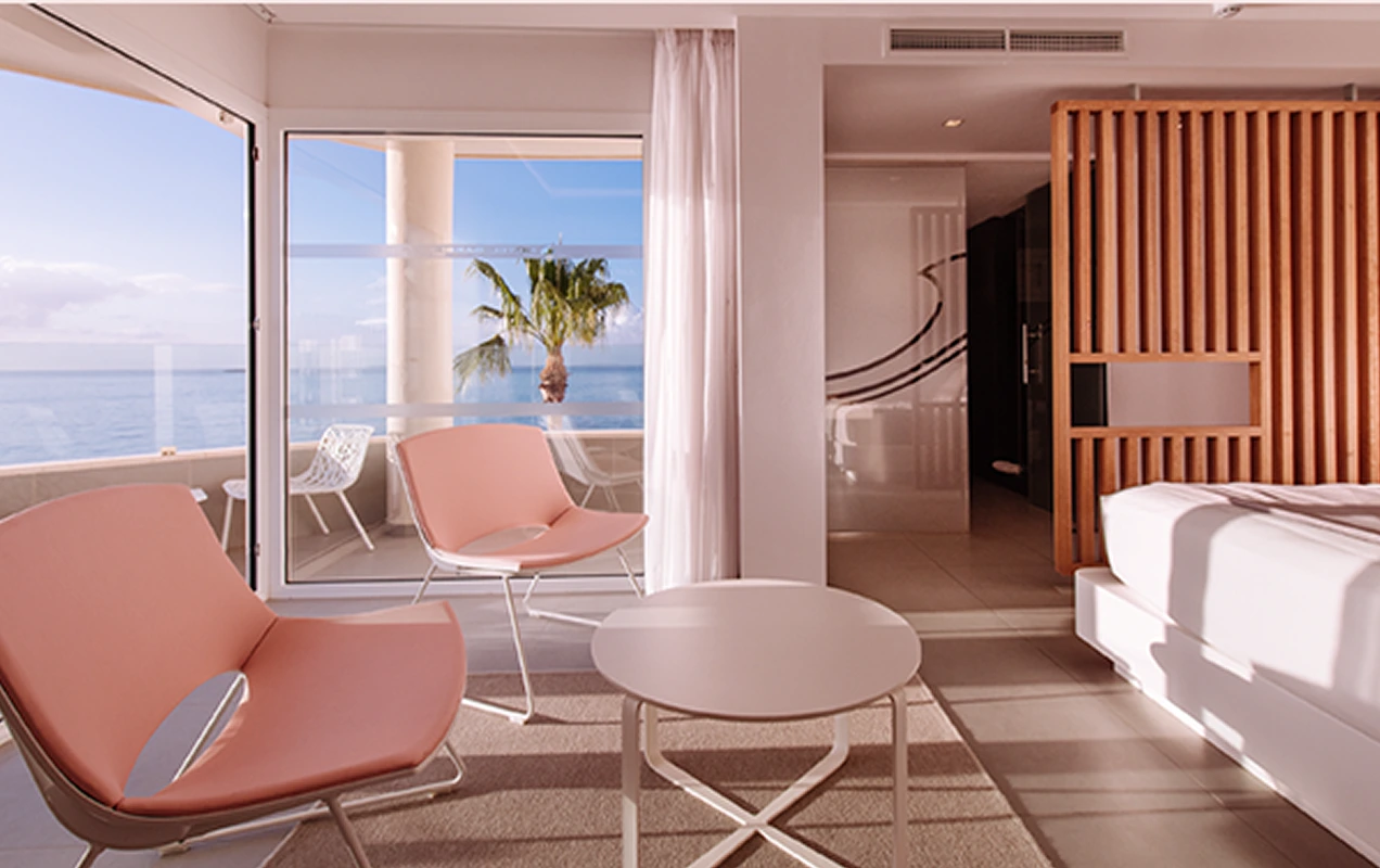 Modern hotel room with two pink chairs, a white round table, a bed, and a balcony overlooking the ocean and a palm tree.