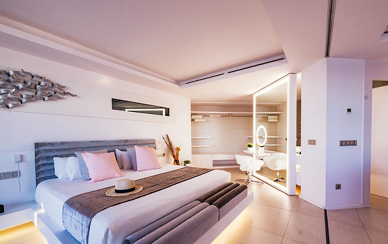 Modern bedroom with a large bed featuring pink and white pillows, a gray headboard, and a hat on the bedspread, illuminated mirror, and white chairs.