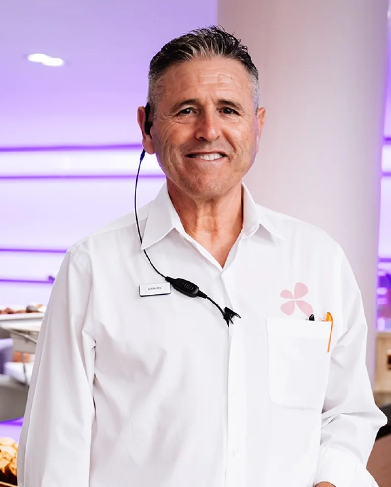 Smiling middle-aged man in a white shirt with a name tag and an earpiece in a brightly lit room.