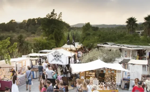 Outdoor market with multiple stalls selling crafts and goods, surrounded by trees and hills in the background.