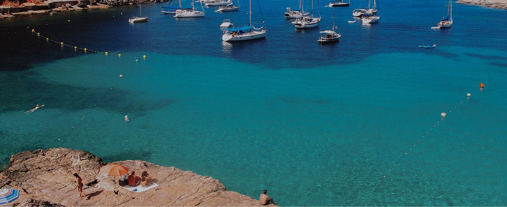 Clear turquoise bay with sailboats anchored offshore and people swimming and relaxing on rocky shore.