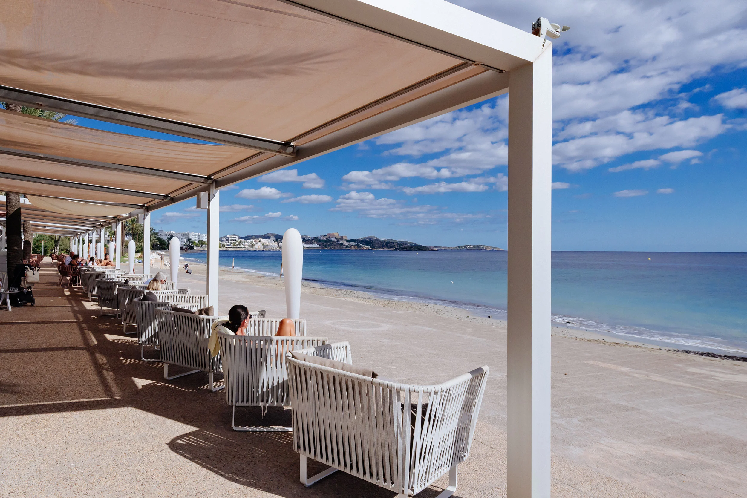 Shaded terrace with white modern chairs facing a sandy beach and calm sea under a blue sky with scattered clouds.