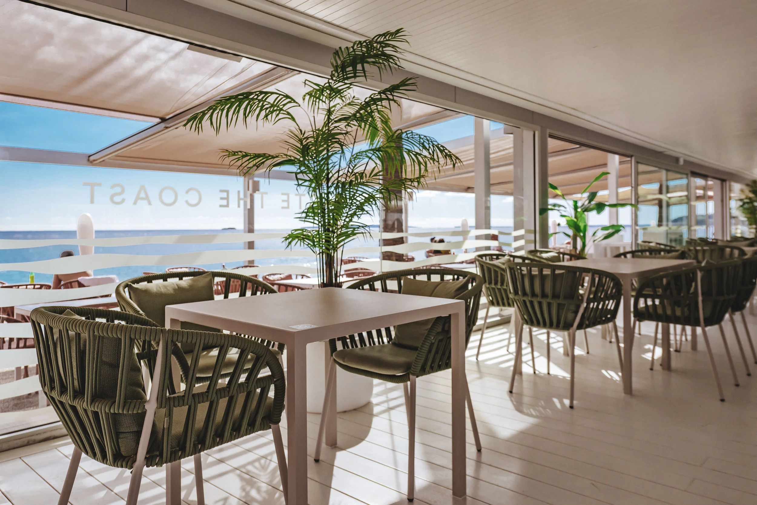 Indoor seating area with tables, green cushioned chairs, and potted plants overlooking a sunny outdoor terrace by the sea.