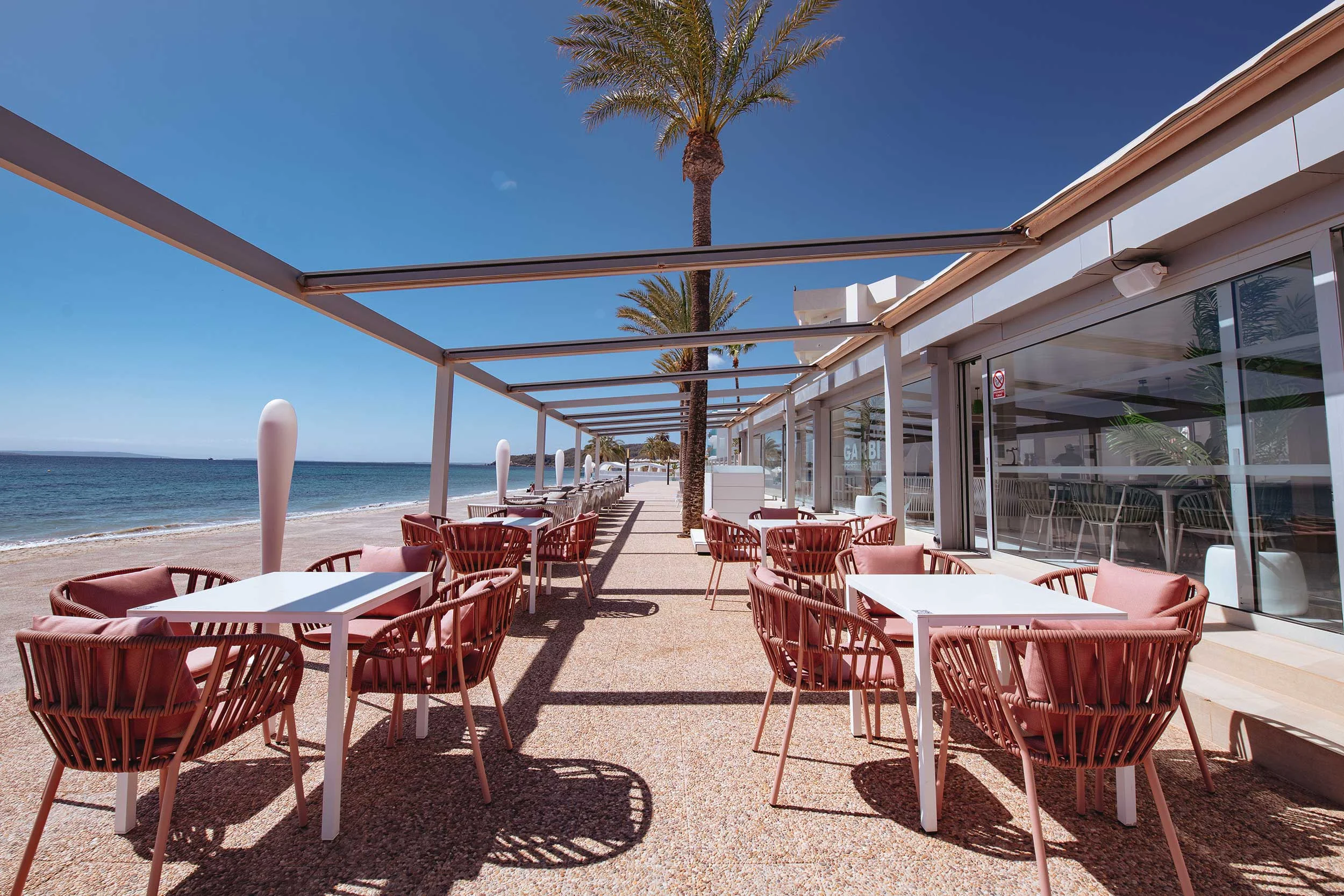Outdoor terrace dining area with white tables and pink cushioned chairs overlooking a beach and ocean under clear blue sky.