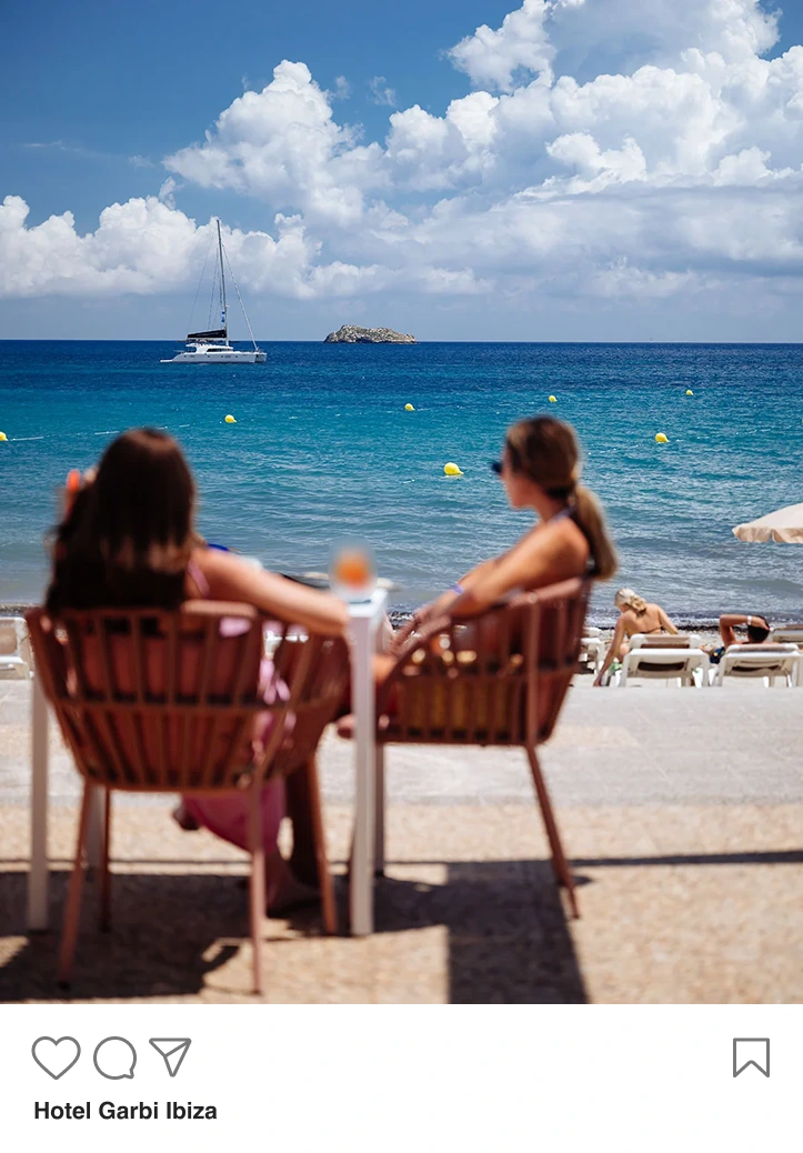 Two women sitting on chairs at a beachside cafe, facing the sea with a sailboat on blue water under a partly cloudy sky.
