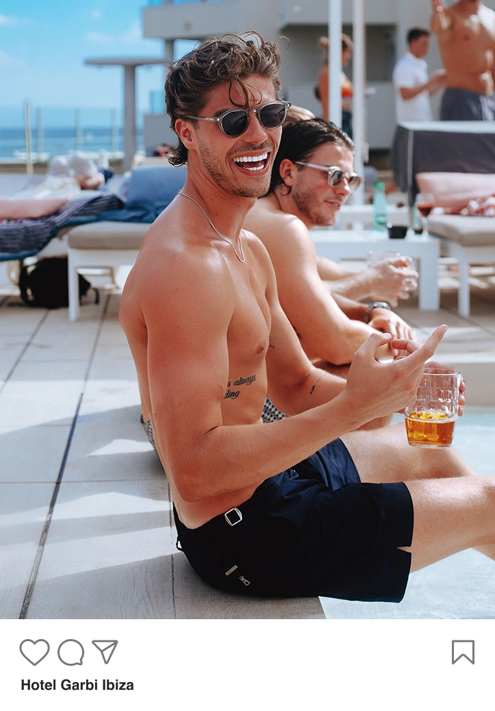 Two shirtless men wearing sunglasses sitting poolside, one smiling and holding a drink.