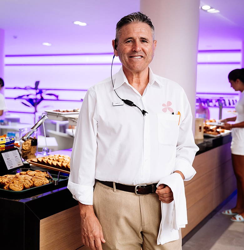 Smiling male waiter in white shirt and beige pants standing in front of pastry display in a restaurant with purple ambient lighting.