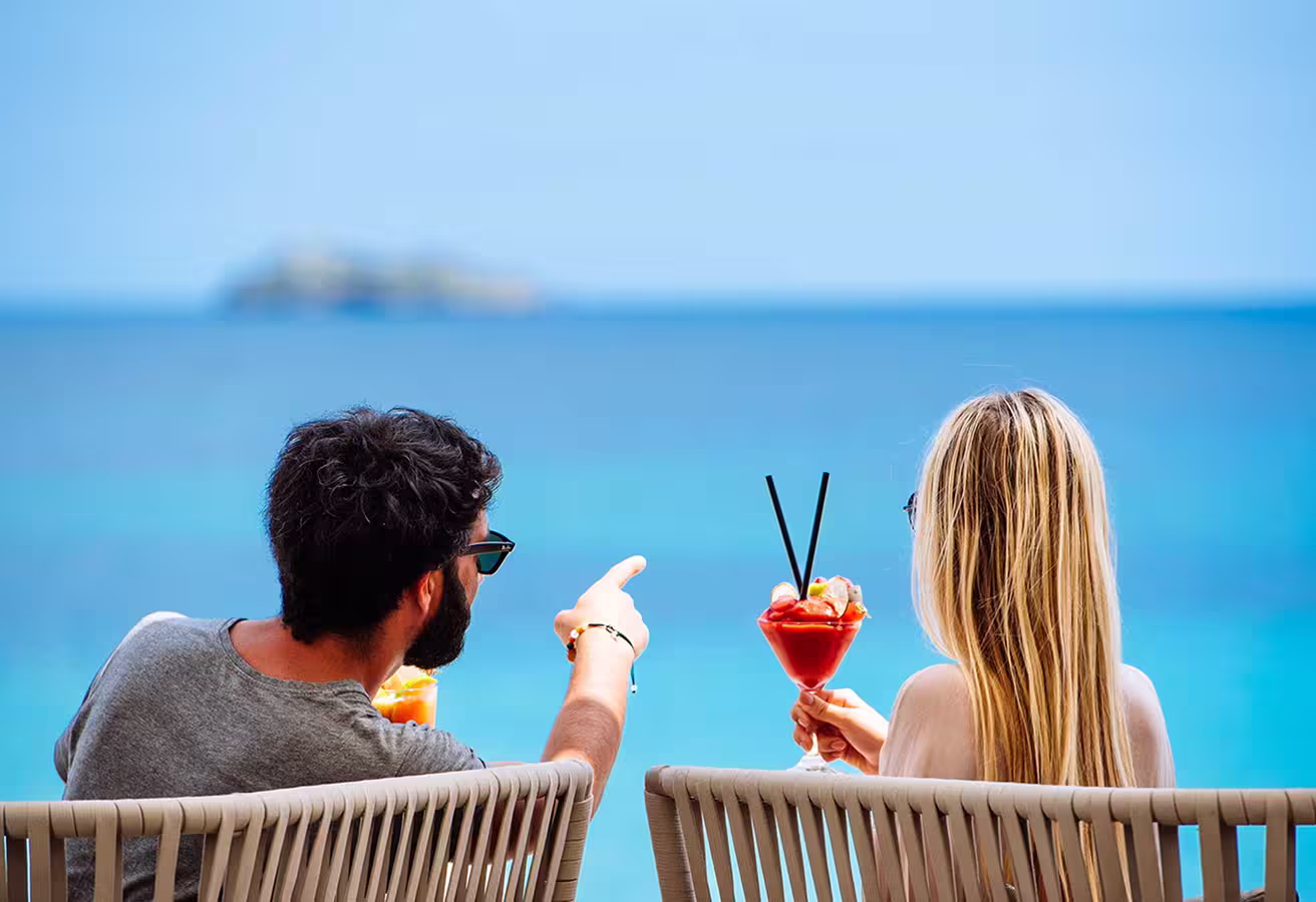 Man and woman sitting outdoors by the sea, clinking colorful cocktails in a celebratory toast.