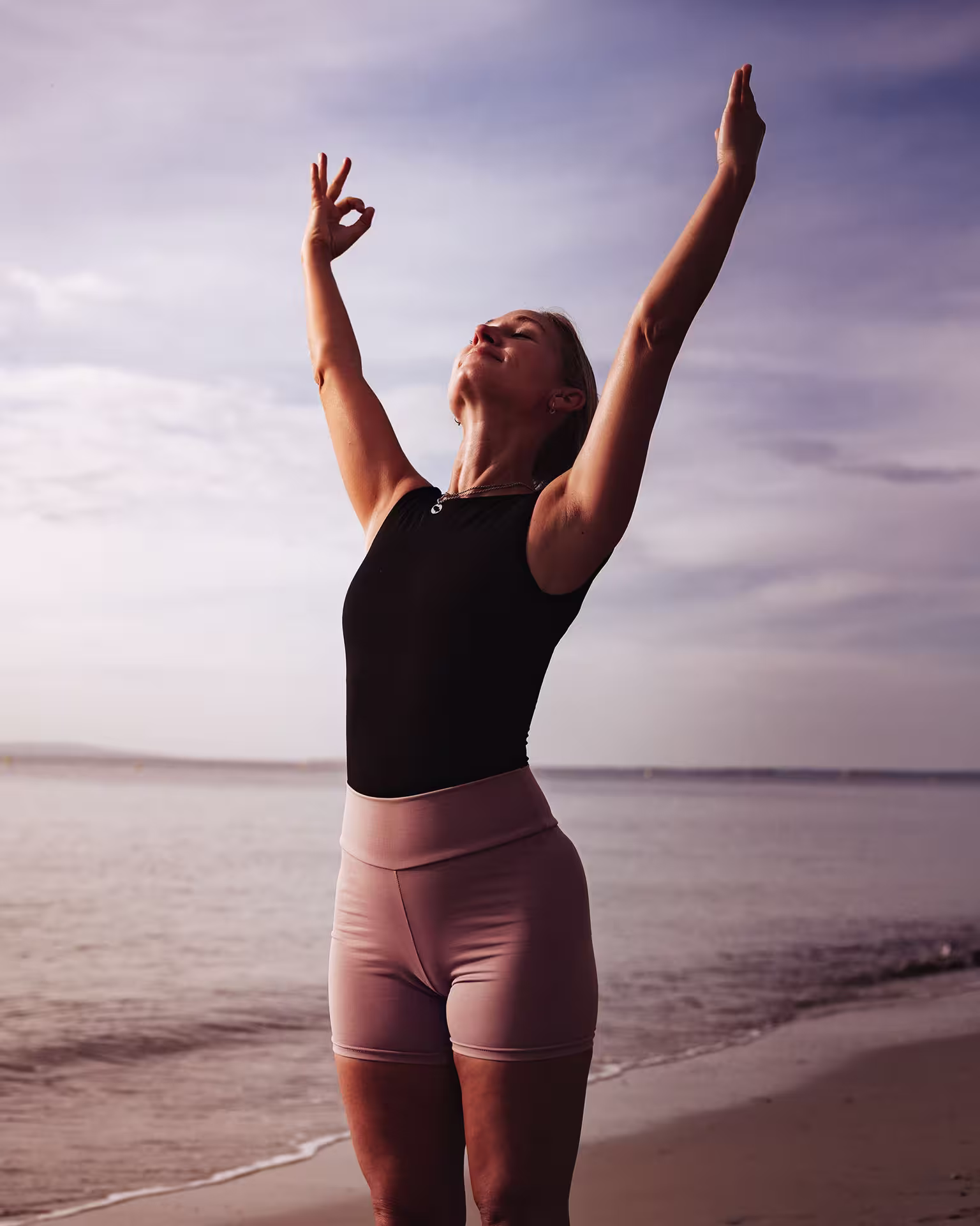 Woman standing on the beach with arms raised, eyes closed, and a serene expression during sunset.
