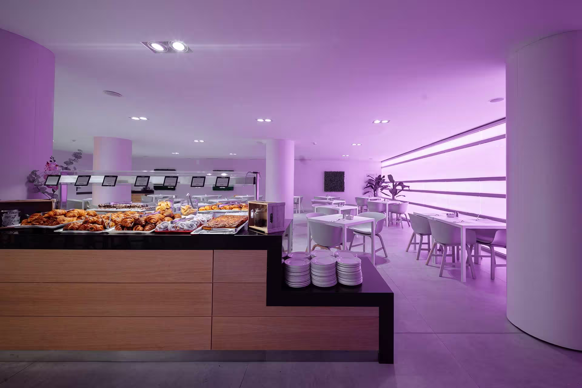 Modern buffet counter with assorted pastries and clean white dining tables under purple lighting.