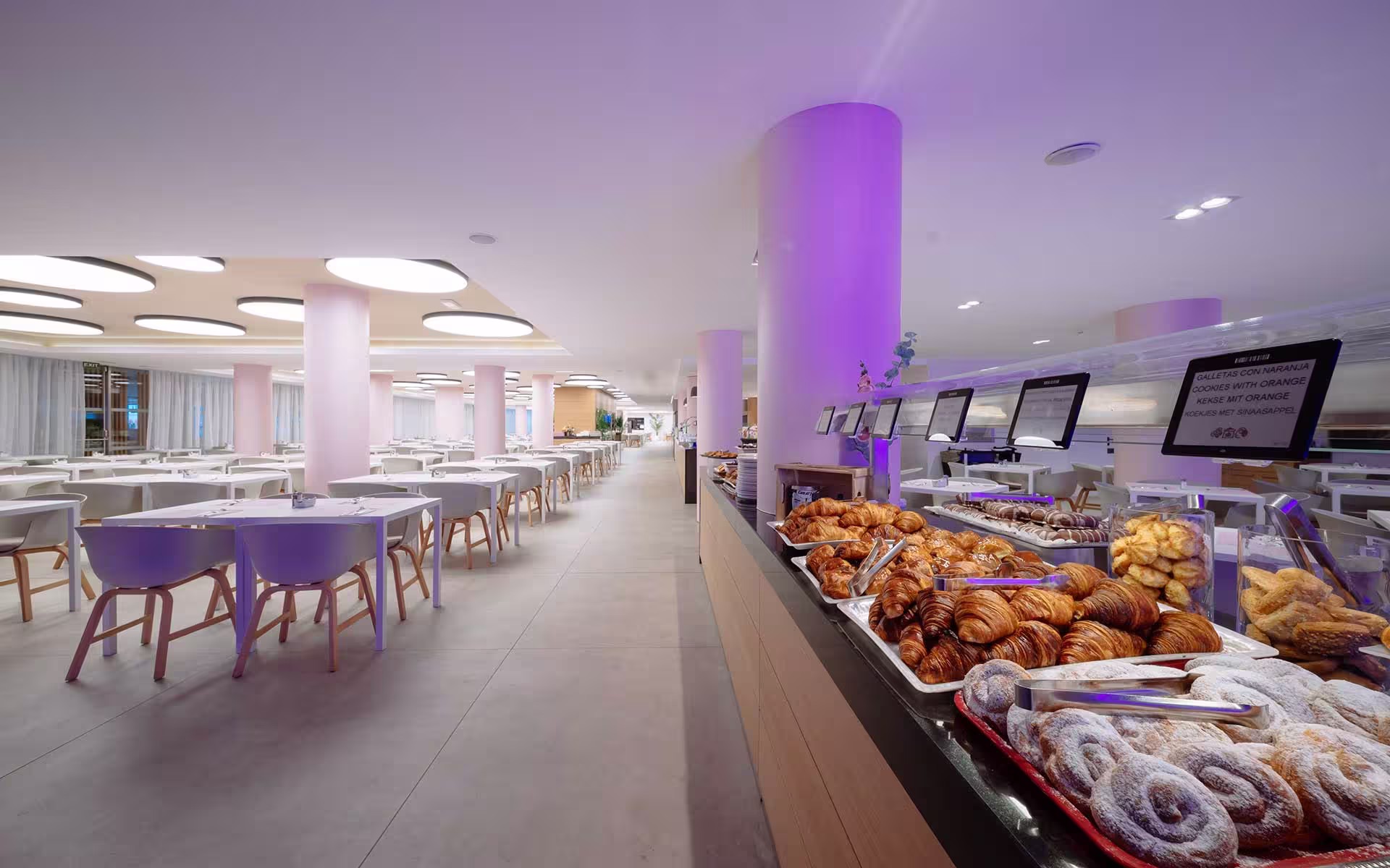 Modern hotel dining area with tables and chairs, and a buffet counter displaying assorted pastries.