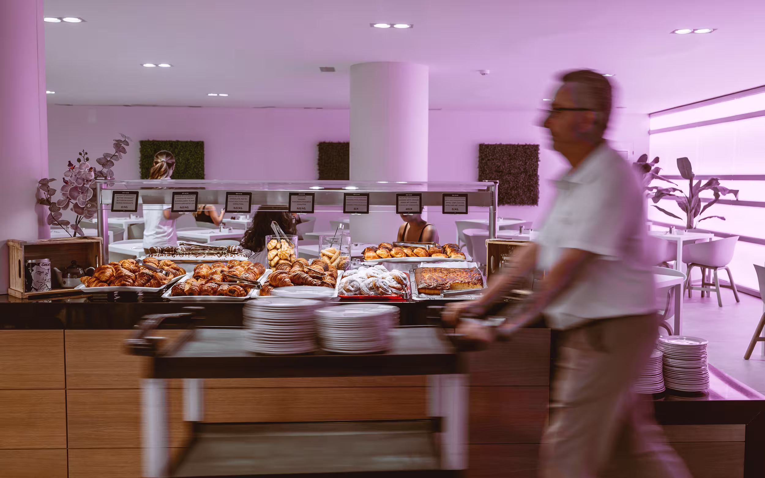 Buffet table with assorted pastries and croissants in a modern cafe with blurred waiter pushing plates on a cart.
