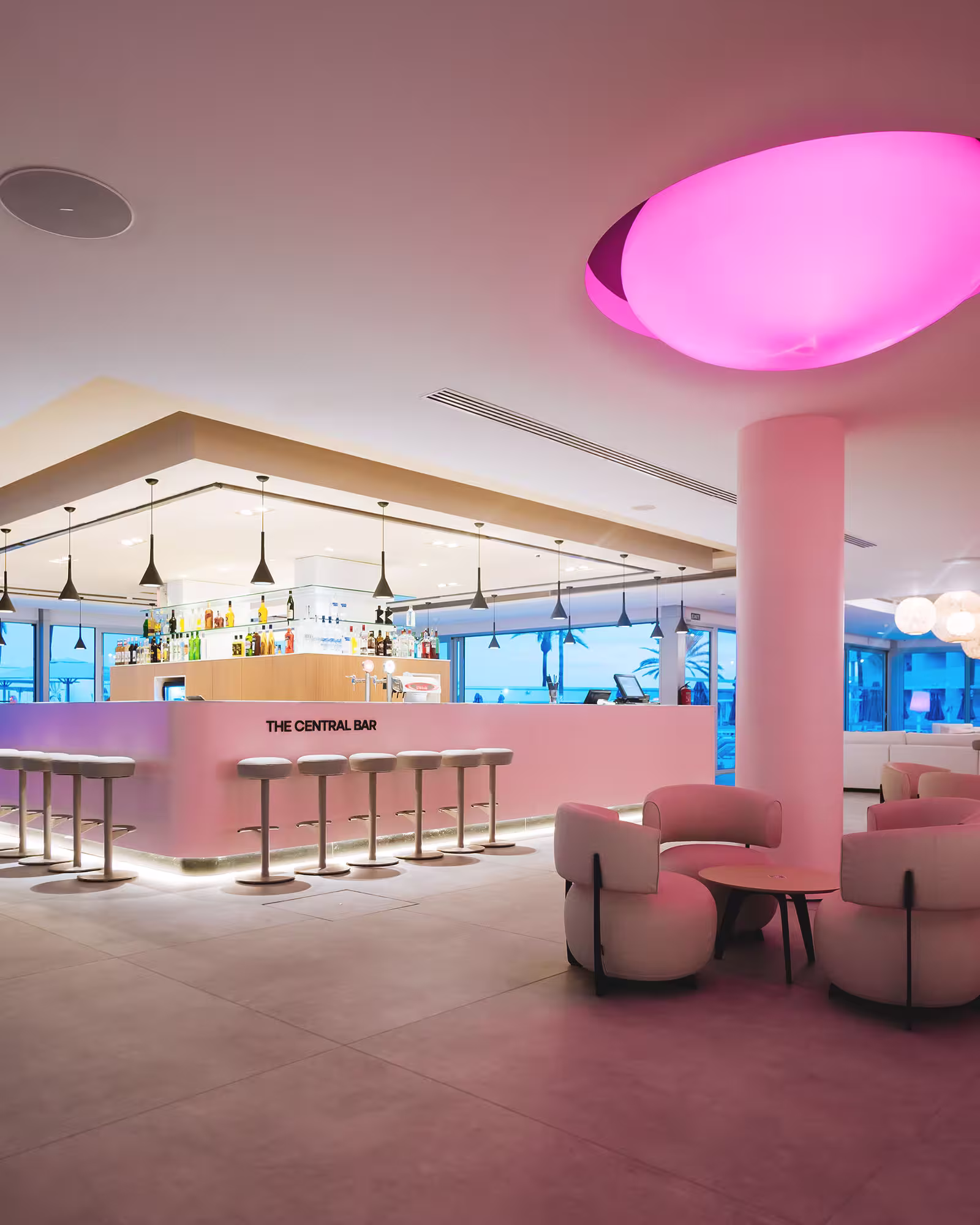 Modern hotel bar interior with stools, circular chairs around tables, and pink ceiling light.