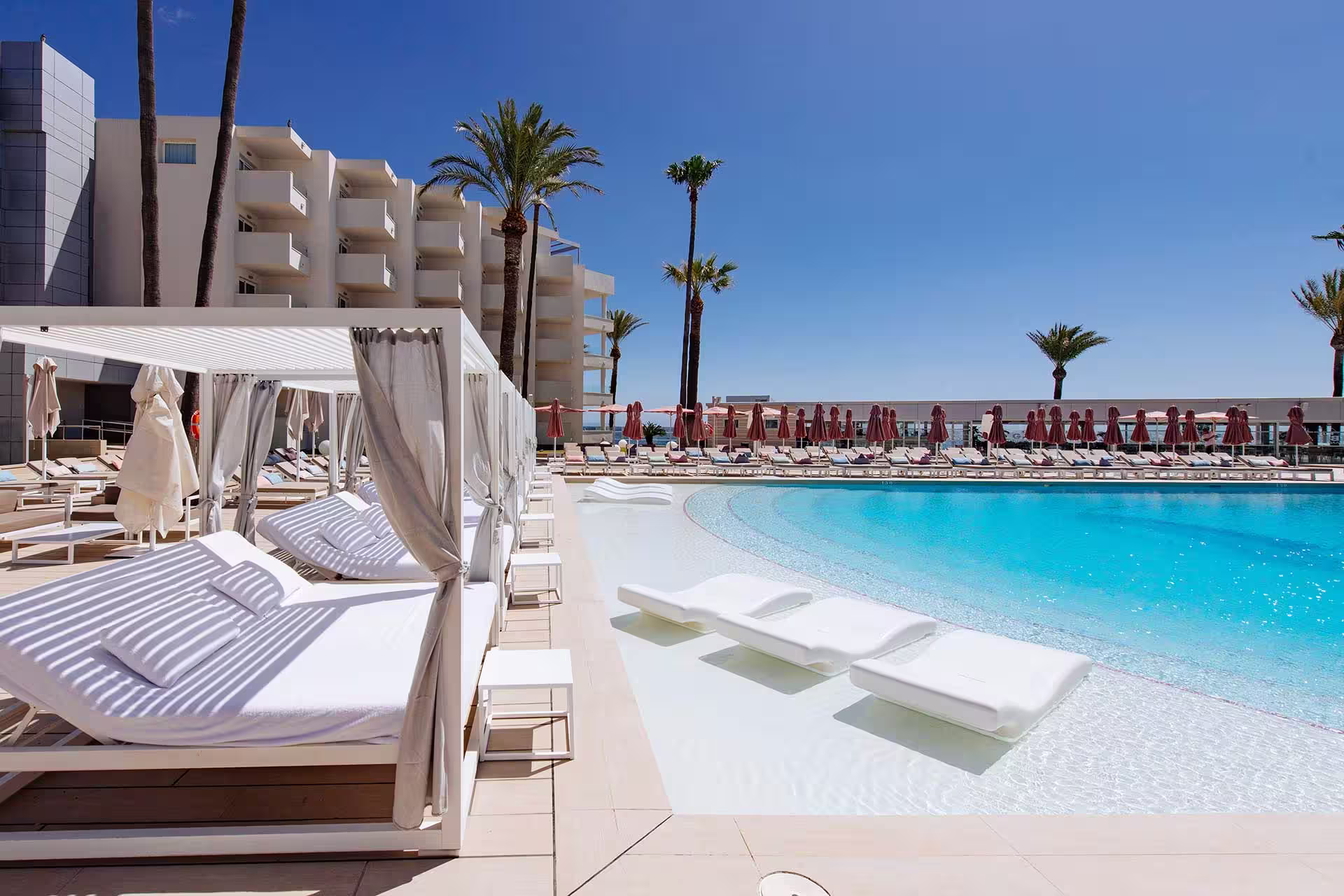 Modern hotel pool area with white sunbeds in shallow water, shaded cabanas, palm trees, and clear blue sky.