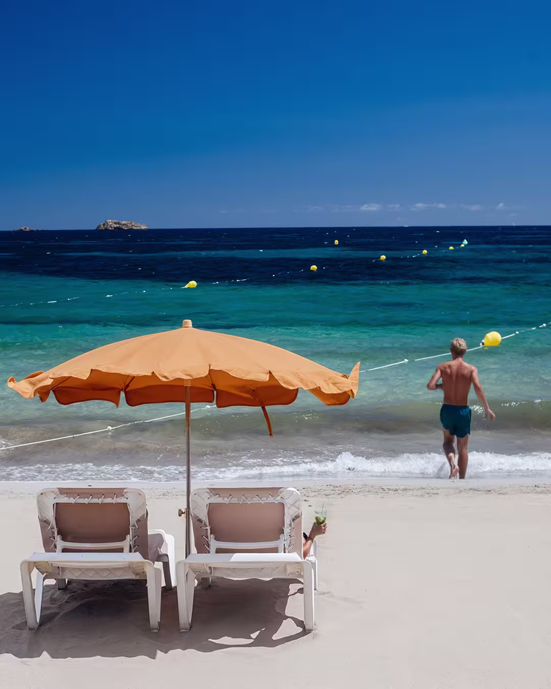 Two empty lounge chairs under an orange beach umbrella with a person walking into the blue-green sea on a sunny beach.