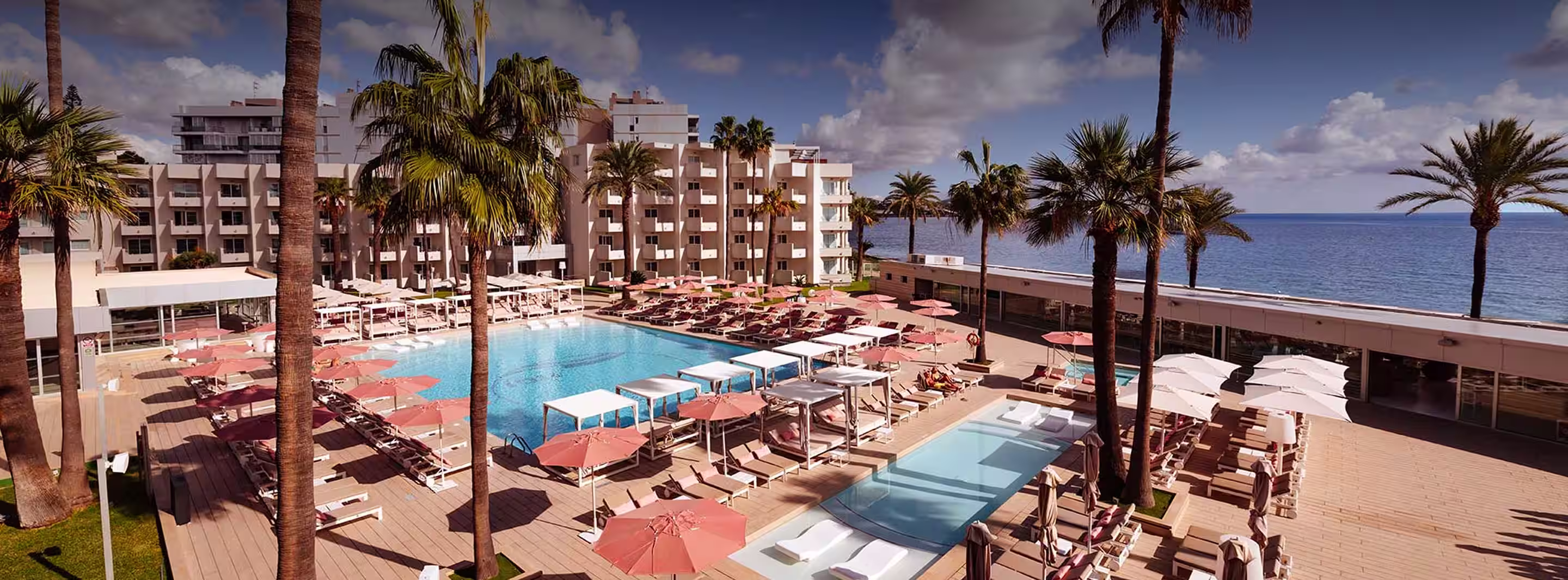 Oceanfront hotel pool area with pink umbrellas, lounge chairs, palm trees, and modern white buildings.